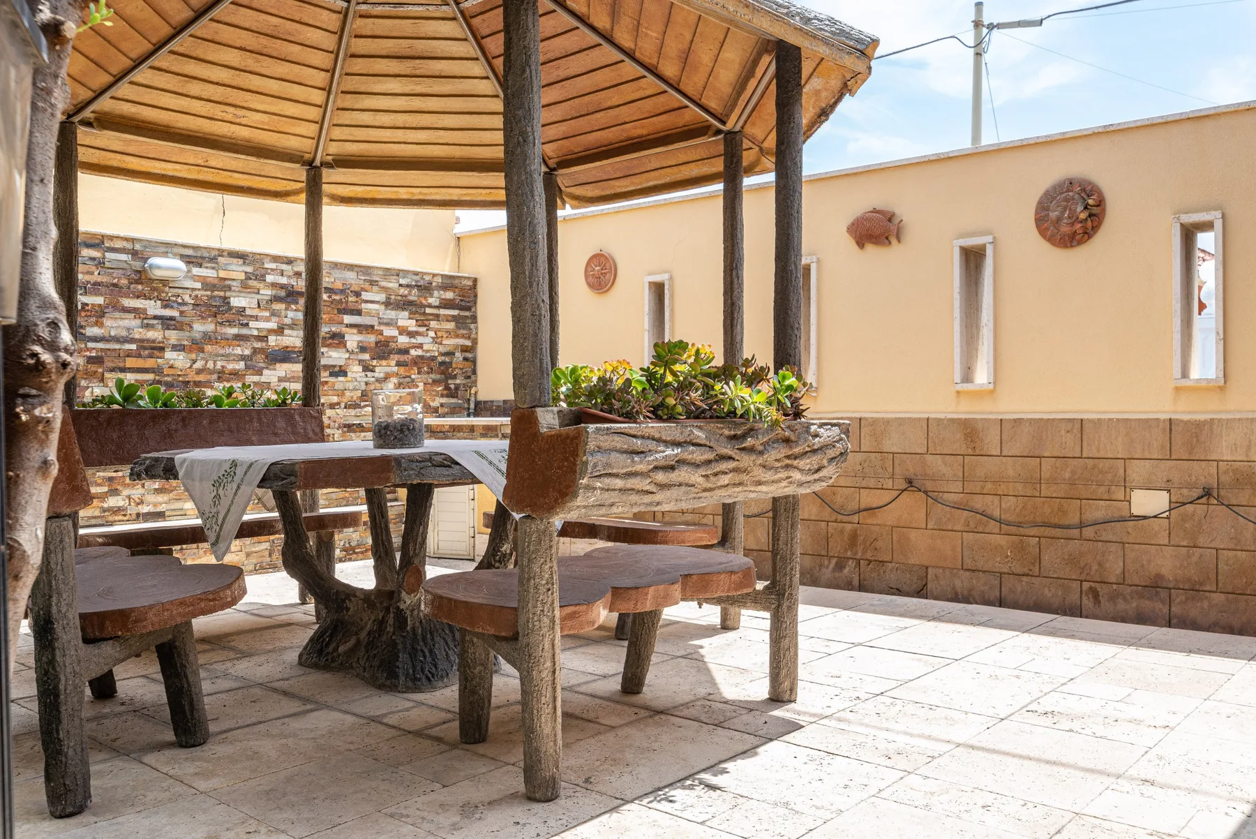 A covered patio with rustic wooden seating and a stone-patterned wall.