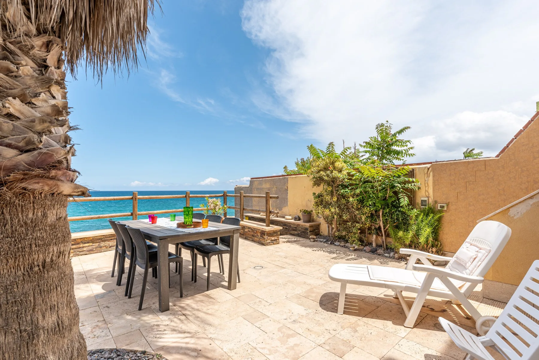 A sunny seaside terrace with outdoor dining furniture and lounge chairs.