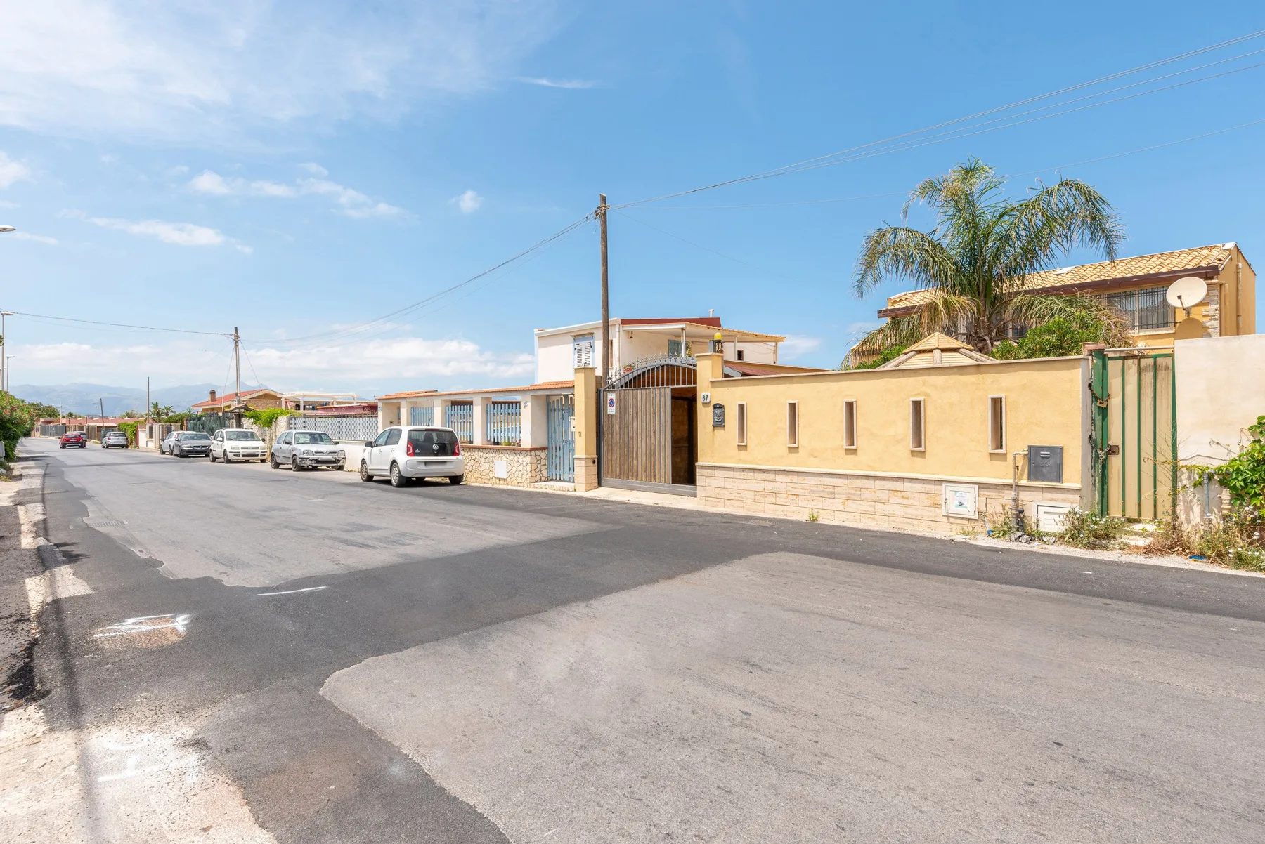 Una calle residencial pasa junto a una propiedad amurallada y varios coches estacionados.