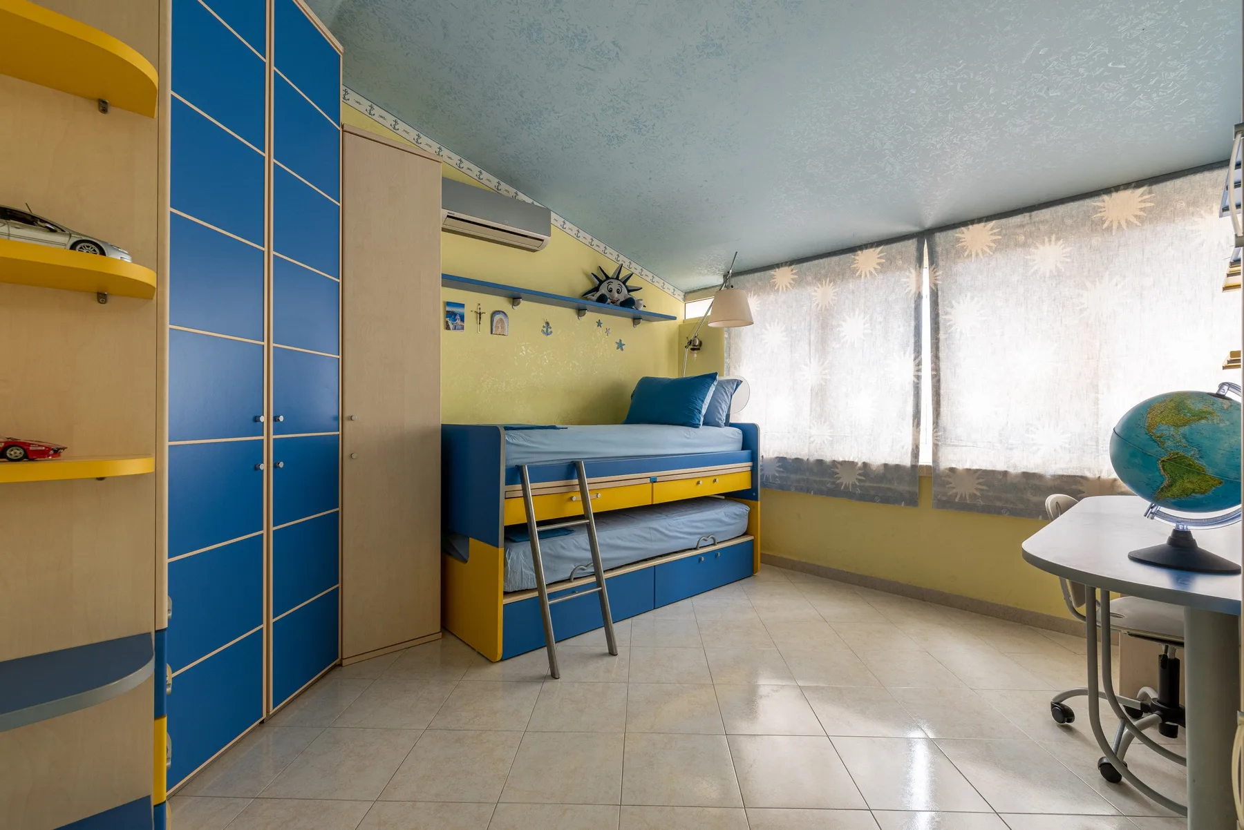 Un dormitorio con una cama azul y amarilla junto a grandes ventanas con cortinas.