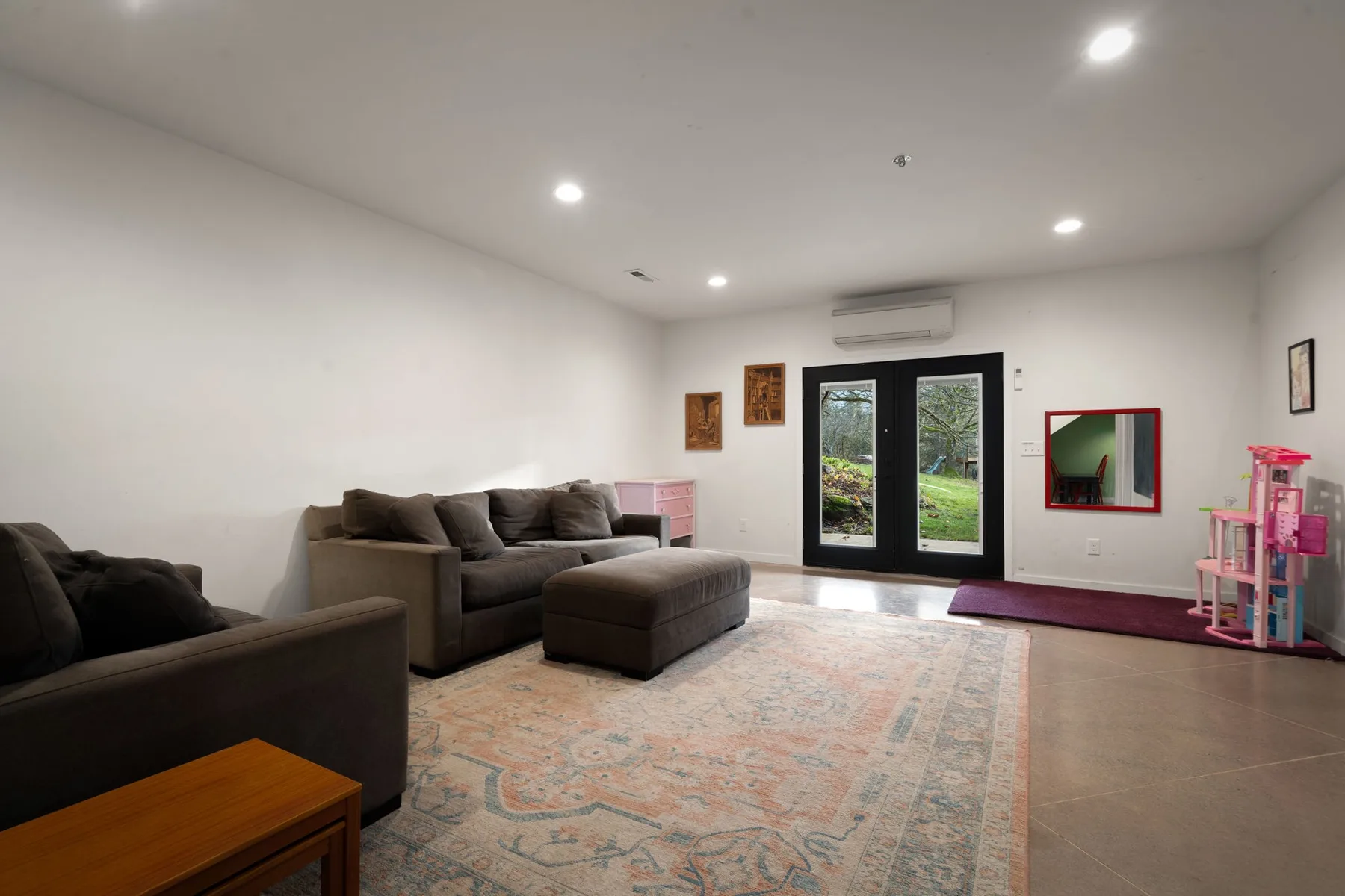 Una sala de estar con asientos oscuros, una alfombra estampada y puertas de vidrio hacia un jardín.