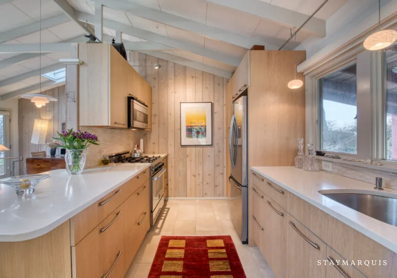 Una cocina luminosa con gabinetes de madera clara, encimeras blancas y una alfombra roja.