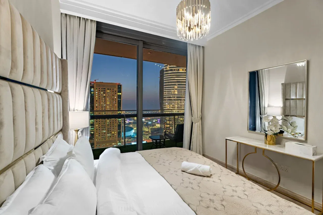 A bedroom with a large bed beside floor-to-ceiling windows overlooking city lights.