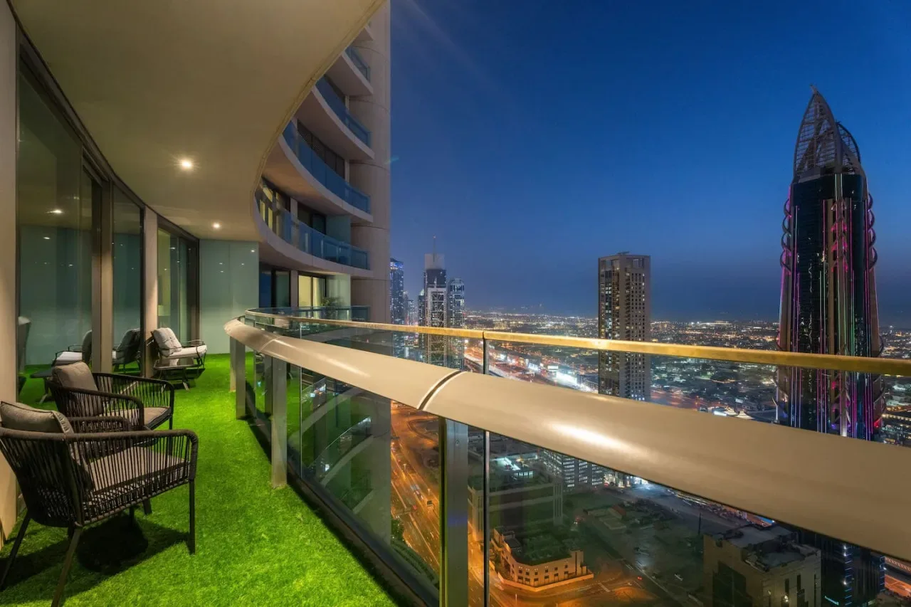 A high-rise balcony overlooks a brightly lit city skyline at dusk.