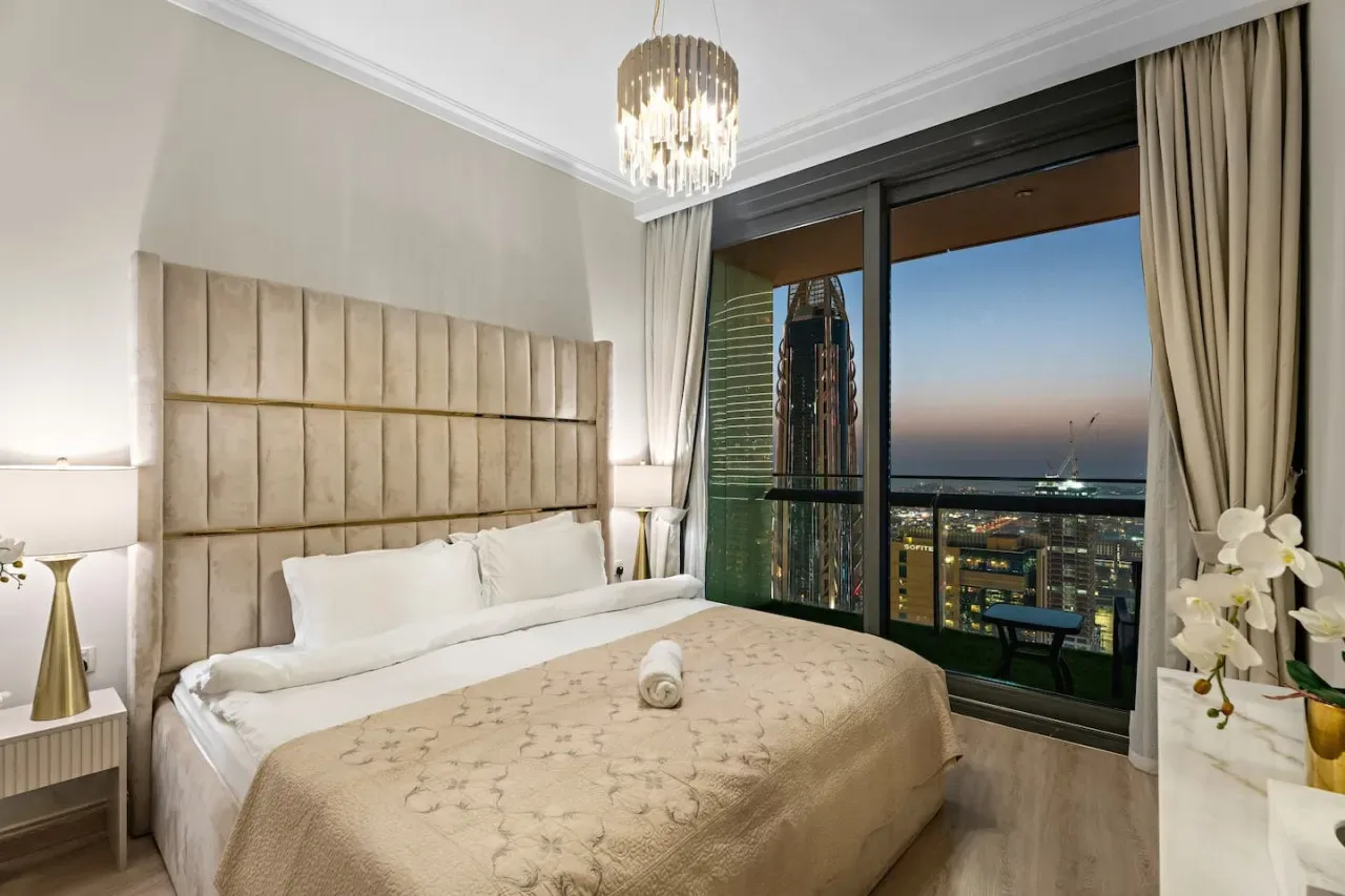 Dormitorio con una cama bien tendida junto a una gran ventana con vistas a las luces de la ciudad.