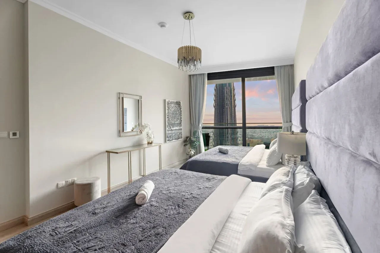 A bright bedroom with two beds, gray bedding, and a large window.