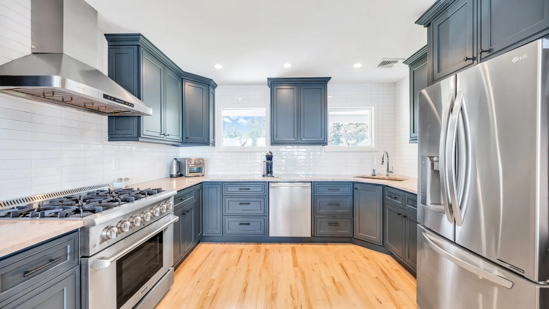 Una cocina luminosa con gabinetes azules, electrodomésticos de acero inoxidable y piso de madera clara.