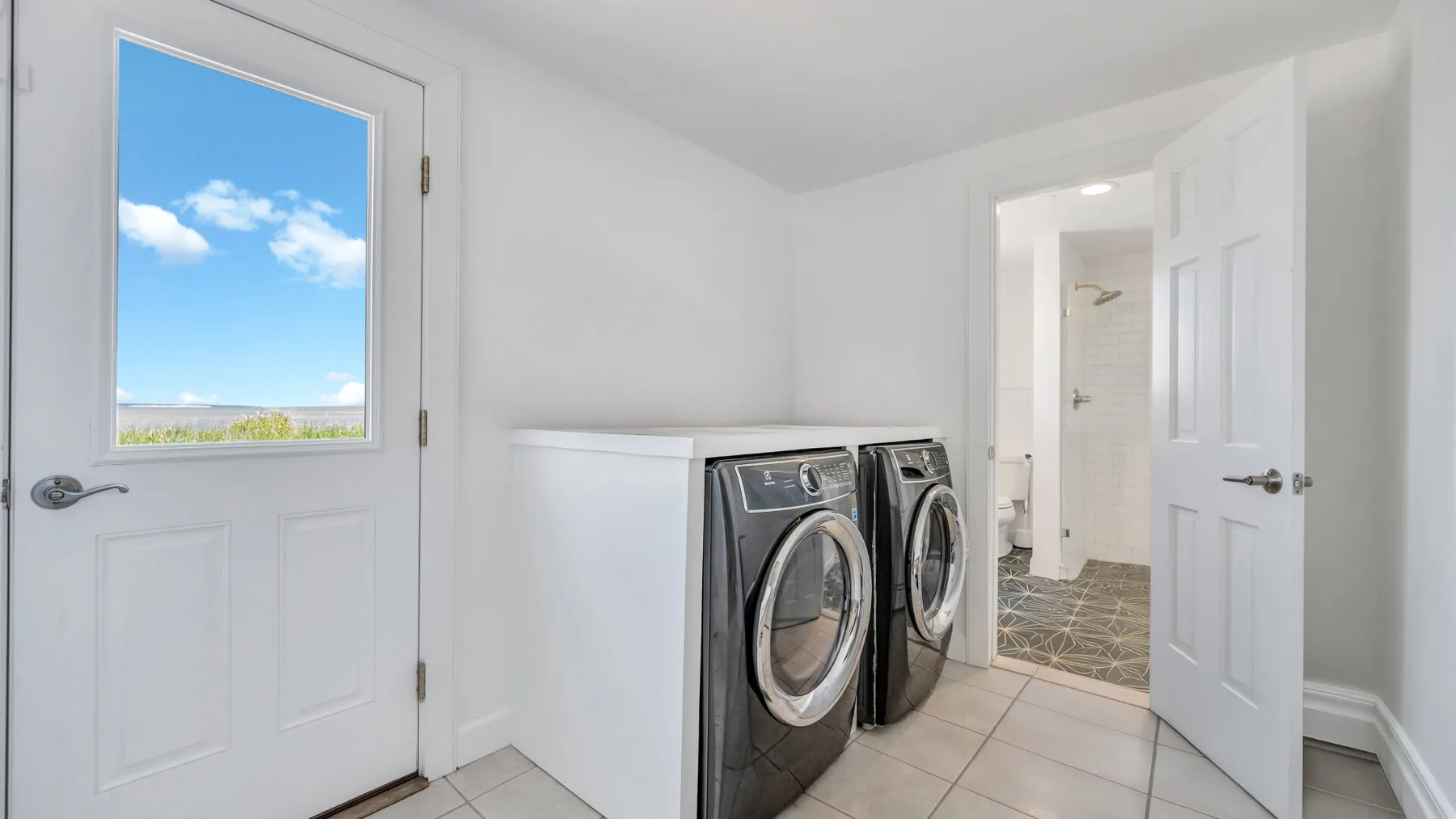 Zona de lavandería con máquinas de carga frontal junto a la entrada abierta de un baño.