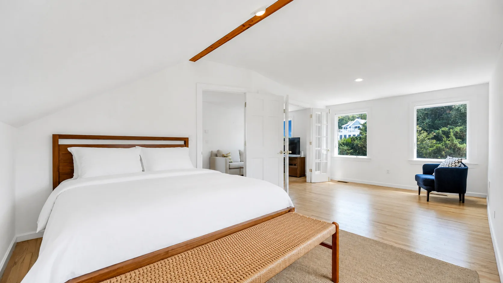 Un dormitorio luminoso con una cama blanca y una zona de estar junto a grandes ventanas.
