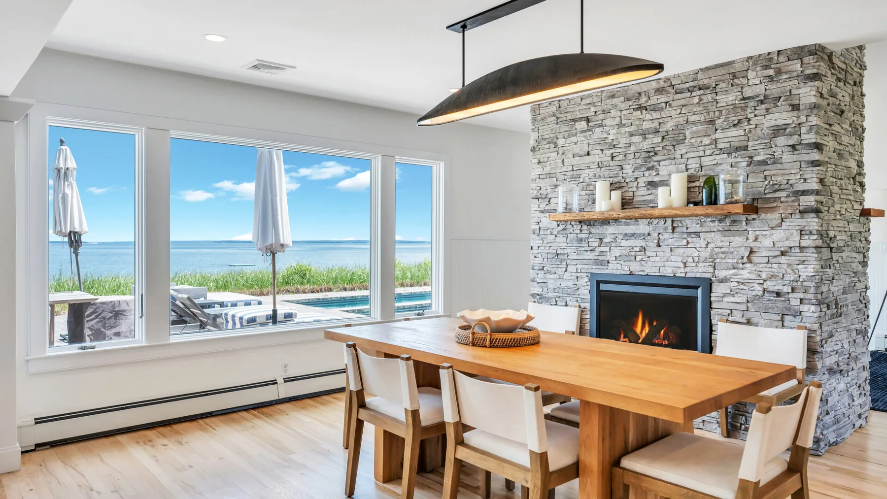 Comedor con una mesa de madera junto a una chimenea de piedra y grandes ventanas.