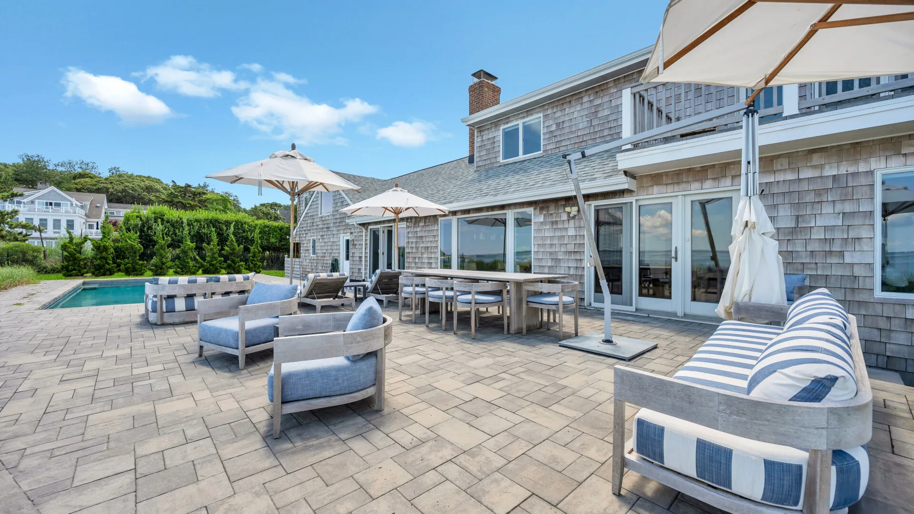 Un patio exterior con asientos, mesas de comedor, sombrillas y una piscina junto a una casa.