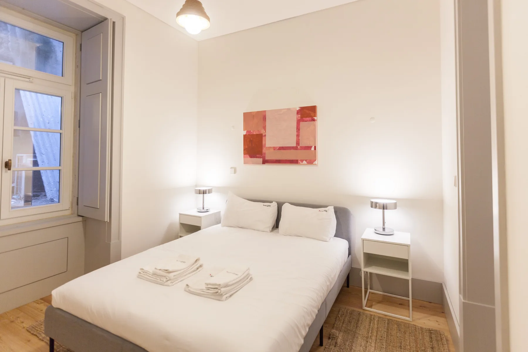 A bright bedroom with a bed, bedside tables, and a large window.