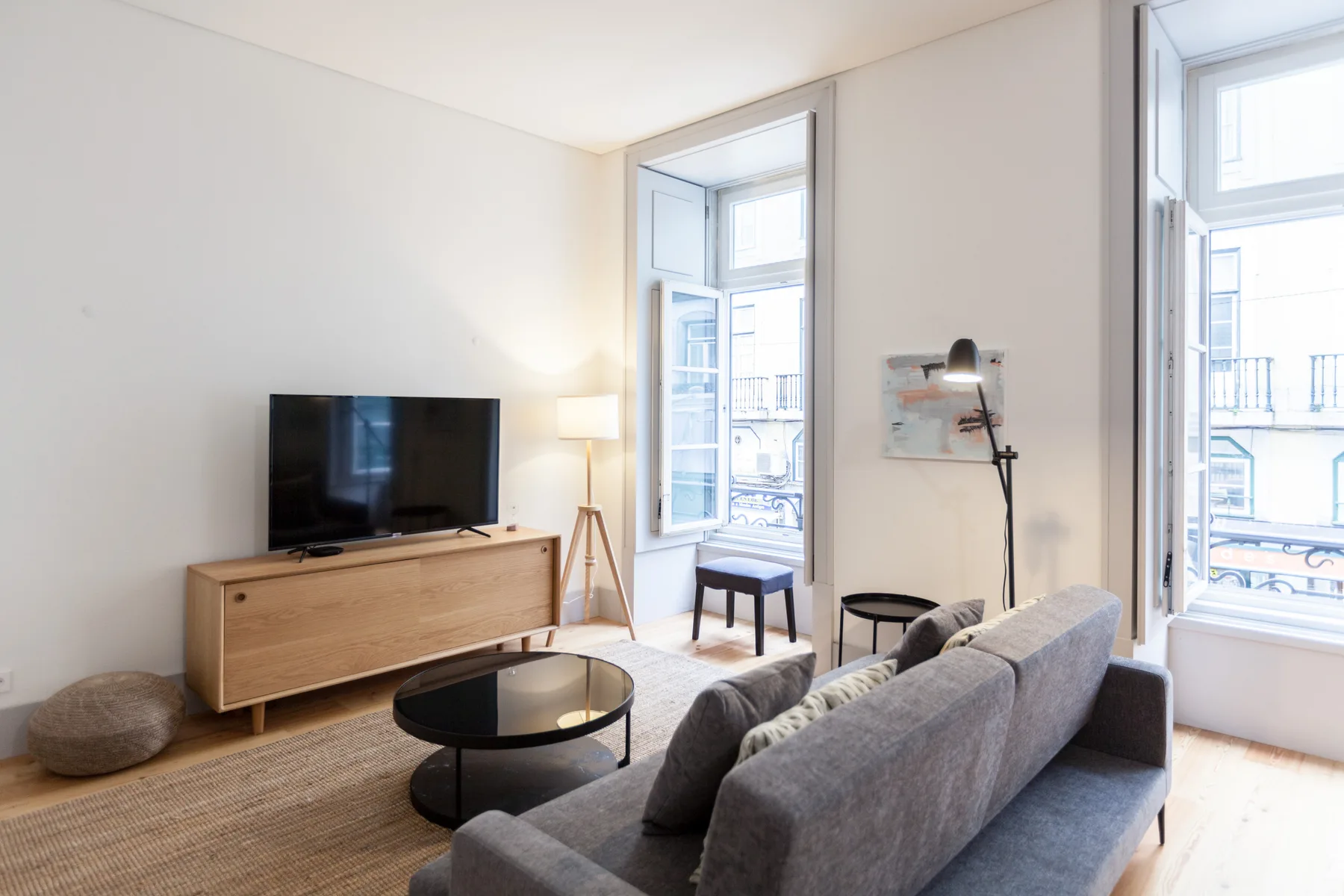 Sala de estar con asientos grises, un televisor y ventanas altas.