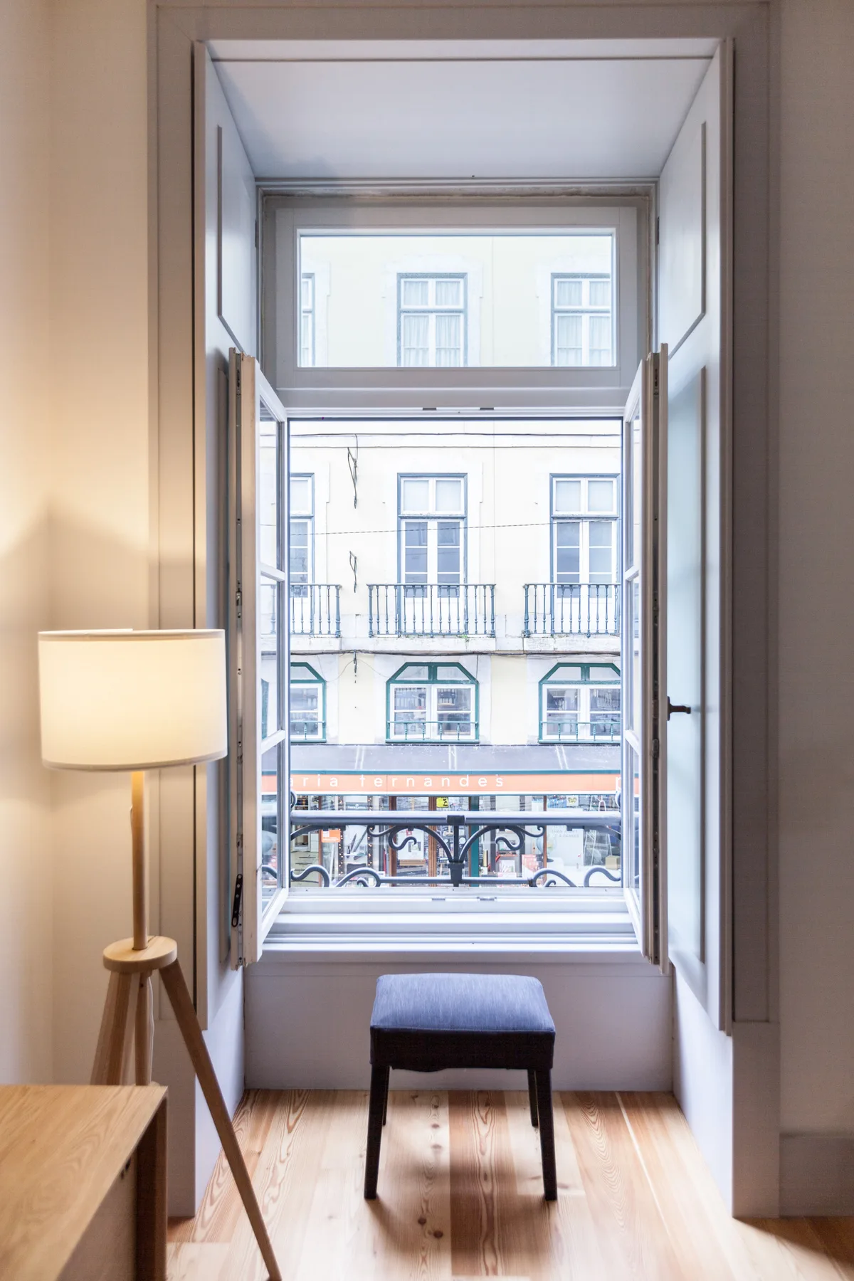 Una habitación luminosa con una ventana abierta, un asiento pequeño y edificios a la vista.