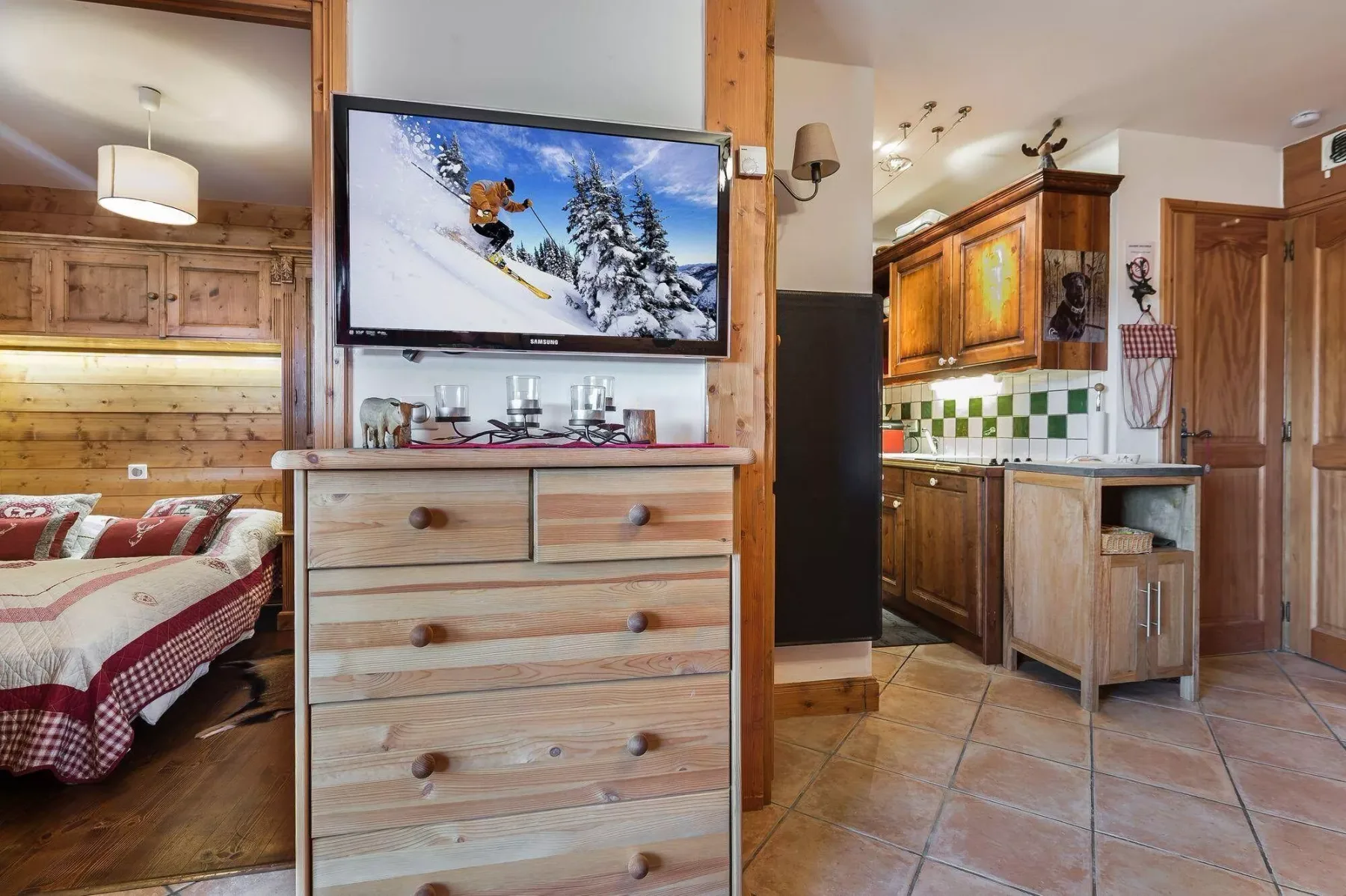 A rustic bedroom and kitchenette are separated by a wooden dresser and television.
