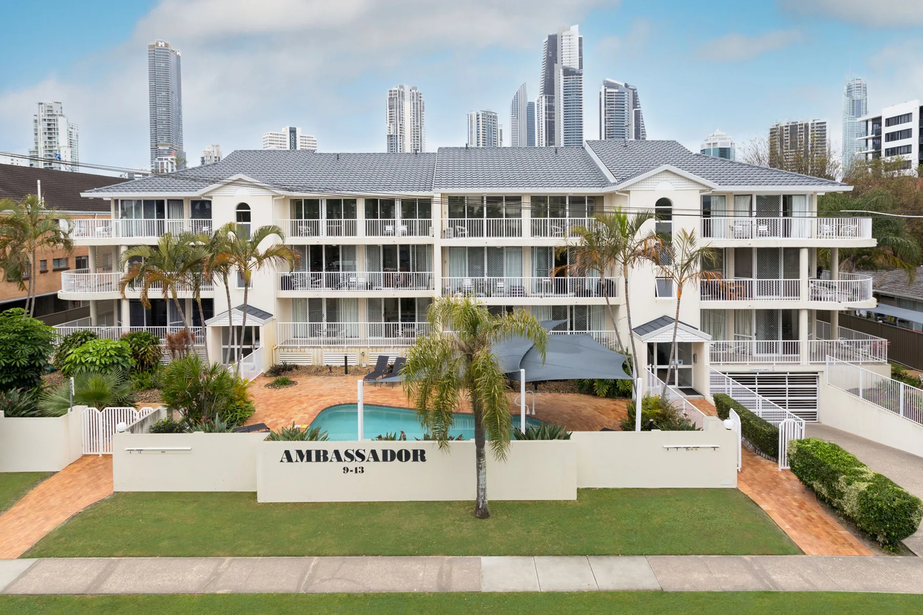 Un edificio de apartamentos de varias plantas se alza entre jardines, con torres urbanas al fondo.