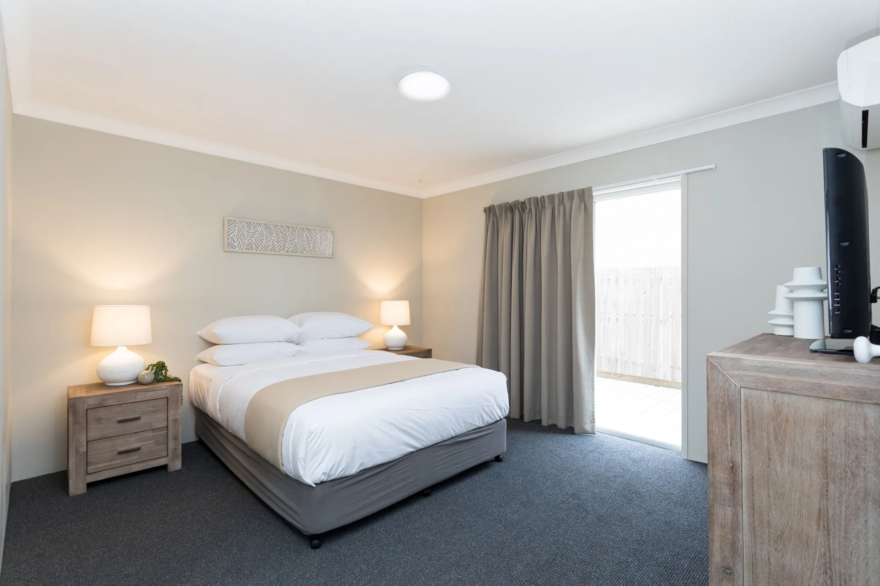 Un dormitorio con una cama tendida, mesitas de noche y una puerta de vidrio con cortinas.