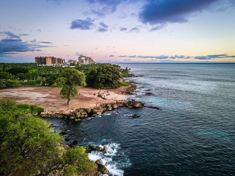 Coastal shoreline with rocky outcrops, greenery, and buildings beneath a cloudy sky.