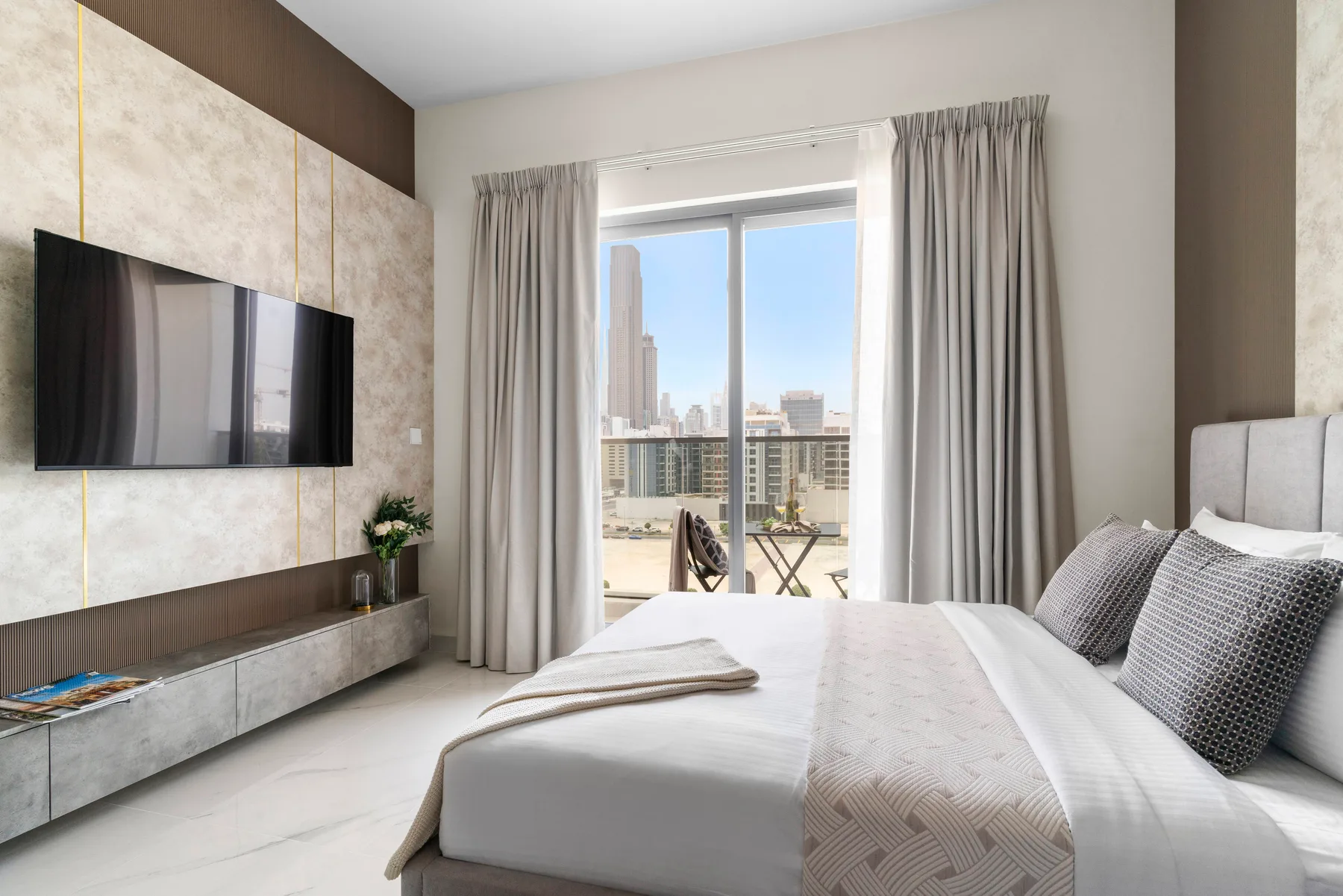 Un dormitorio luminoso con cama, televisor en la pared y una gran ventana con vistas a la ciudad.