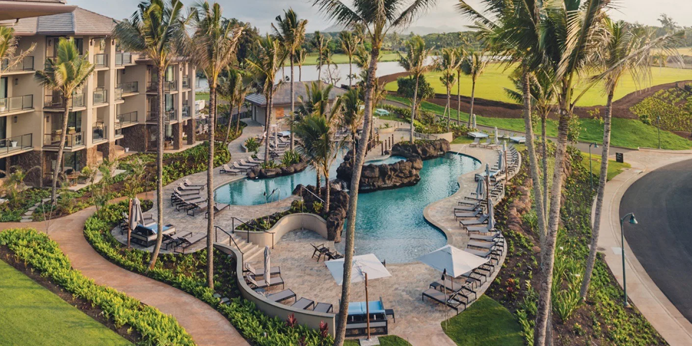 Una piscina de complejo rodeada de palmeras, con jardines y edificios cercanos.