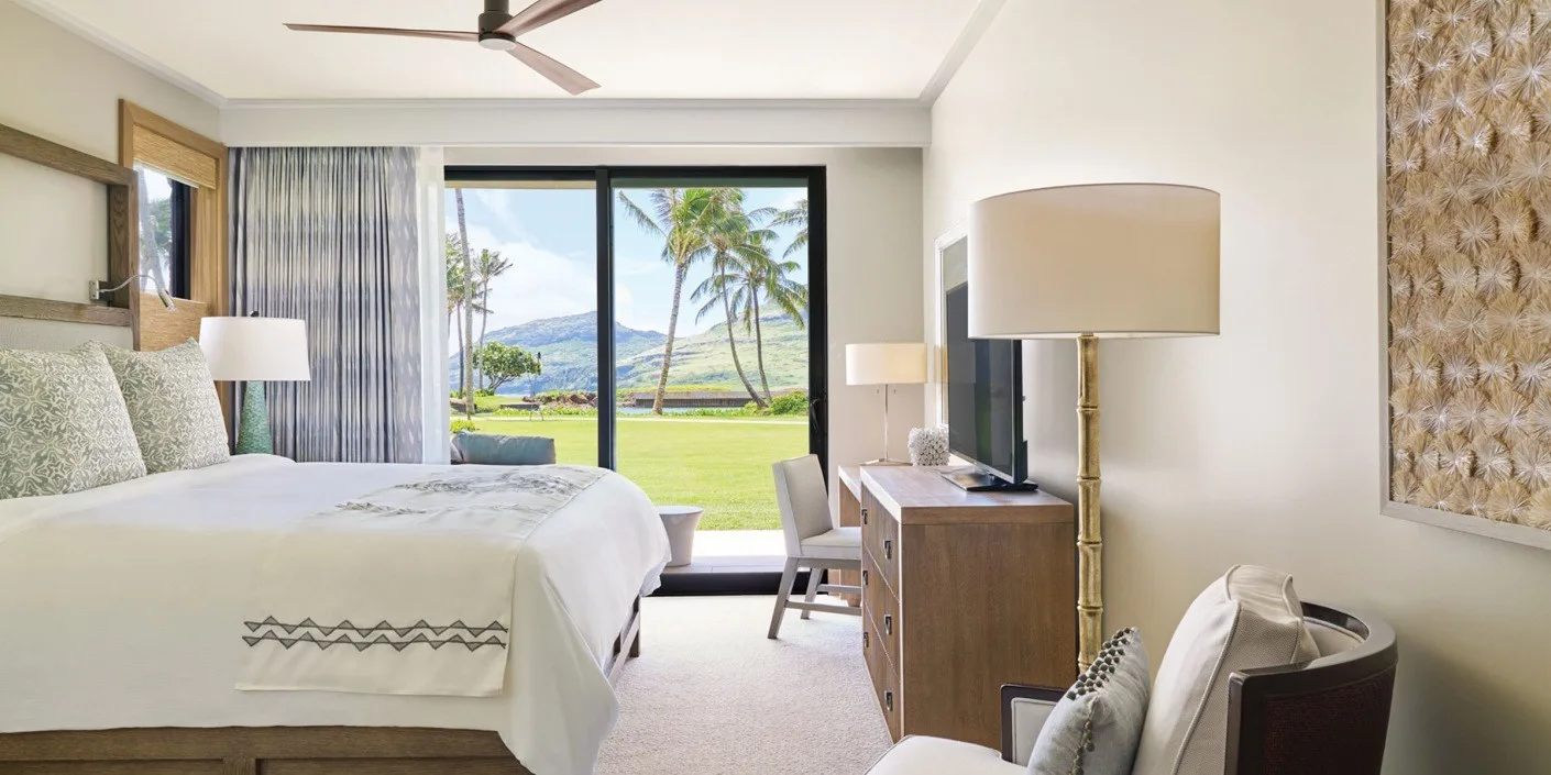 Un dormitorio luminoso tiene una cama grande junto a una ventana con vistas a palmeras y vegetación.