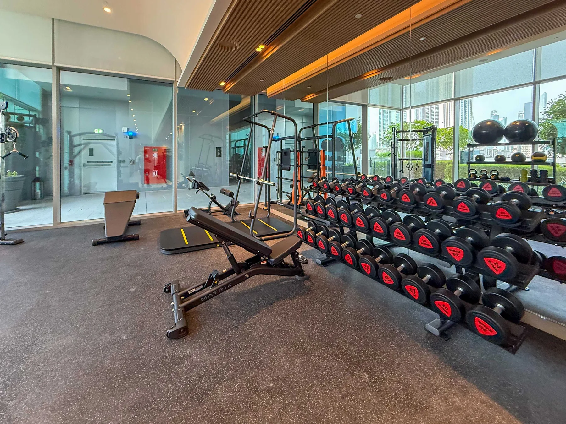 Un gimnasio con estantes de pesas y equipos de ejercicio junto a grandes ventanales.