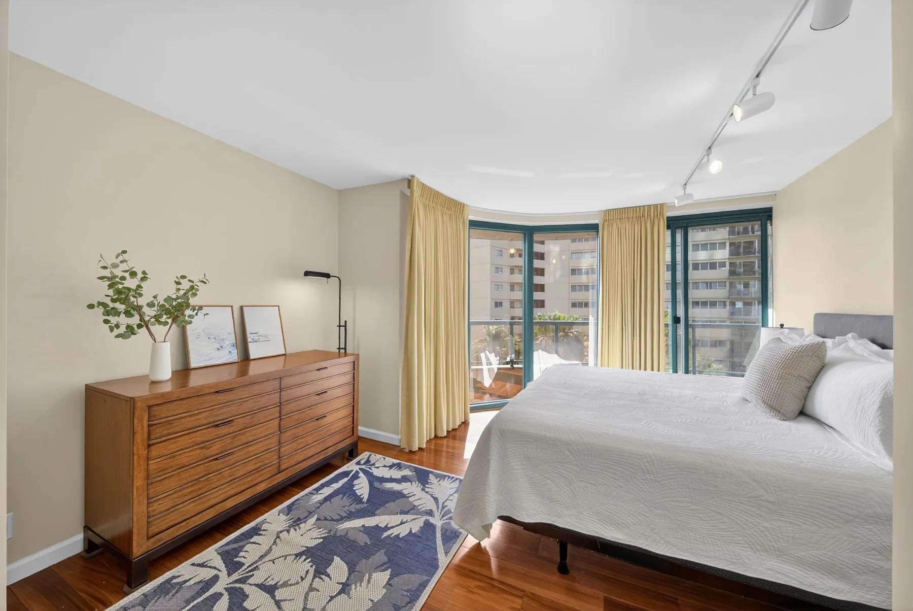 A bedroom with a large bed, wooden dresser, and windows overlooking nearby buildings.
