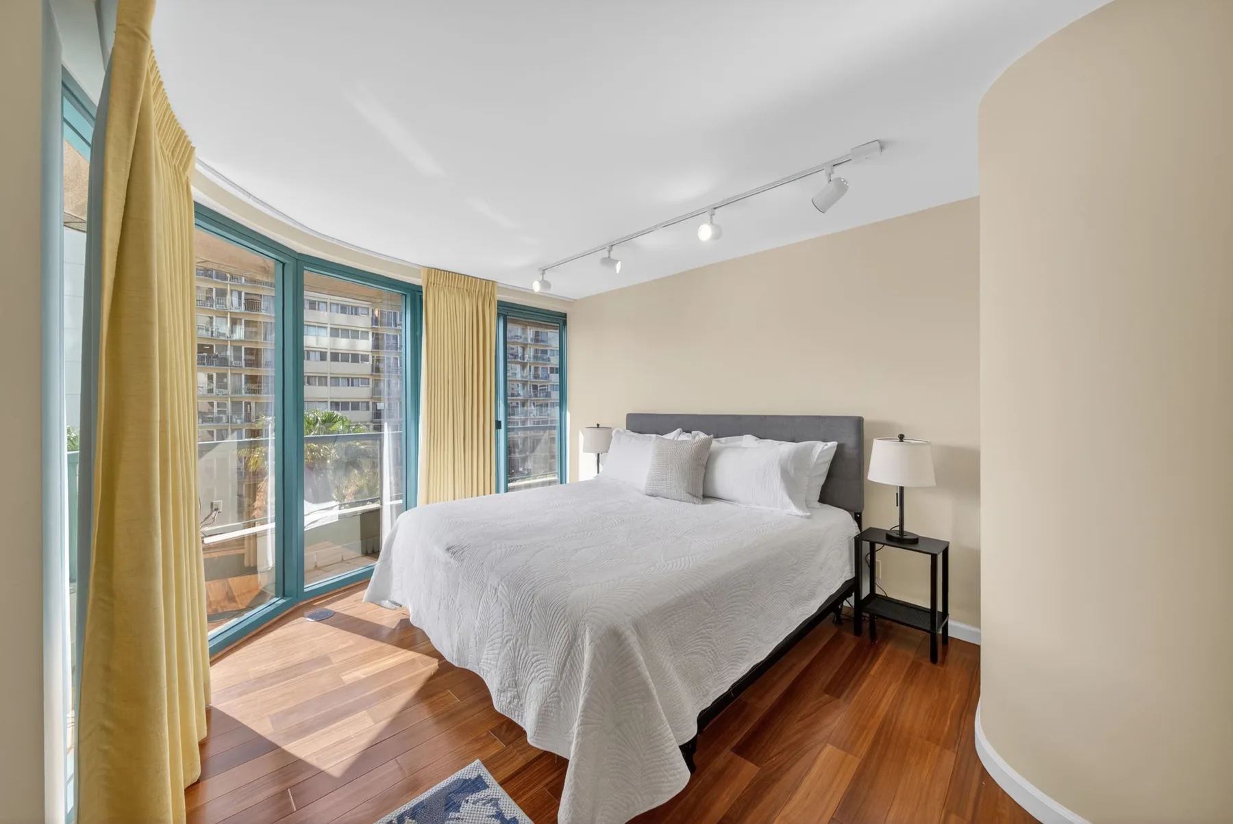 A bright bedroom with a neatly made bed beside large windows and yellow curtains.