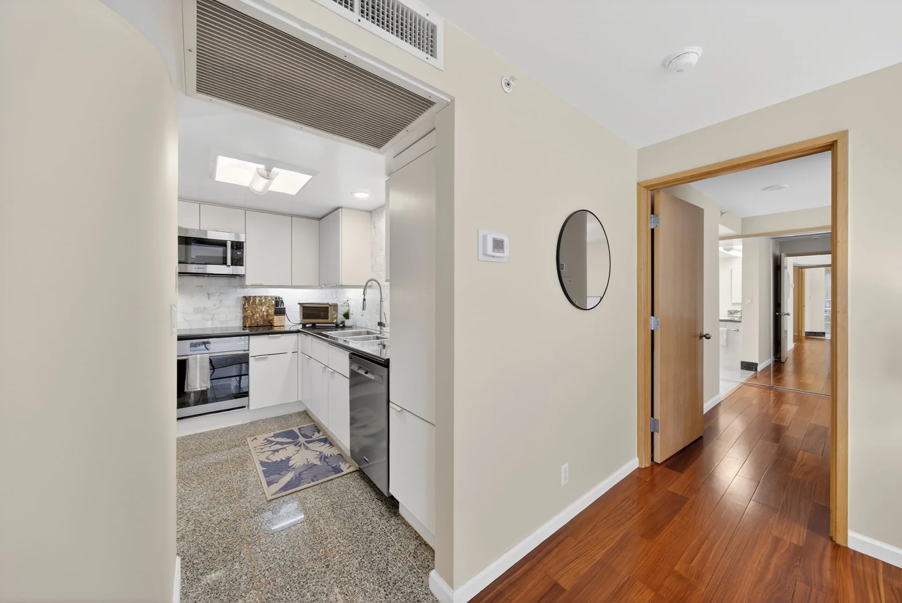 A kitchen opens onto a hallway with wood flooring and a wall mirror.