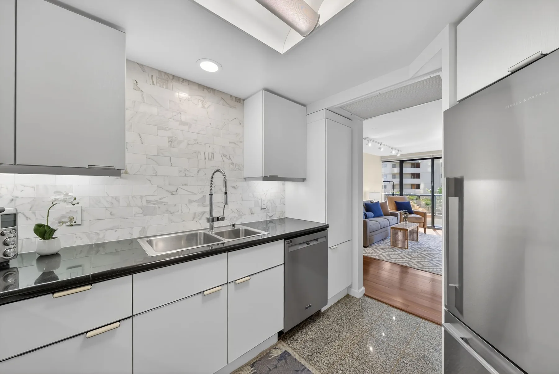 A modern kitchen with light cabinets and a dark countertop opens into a living area.