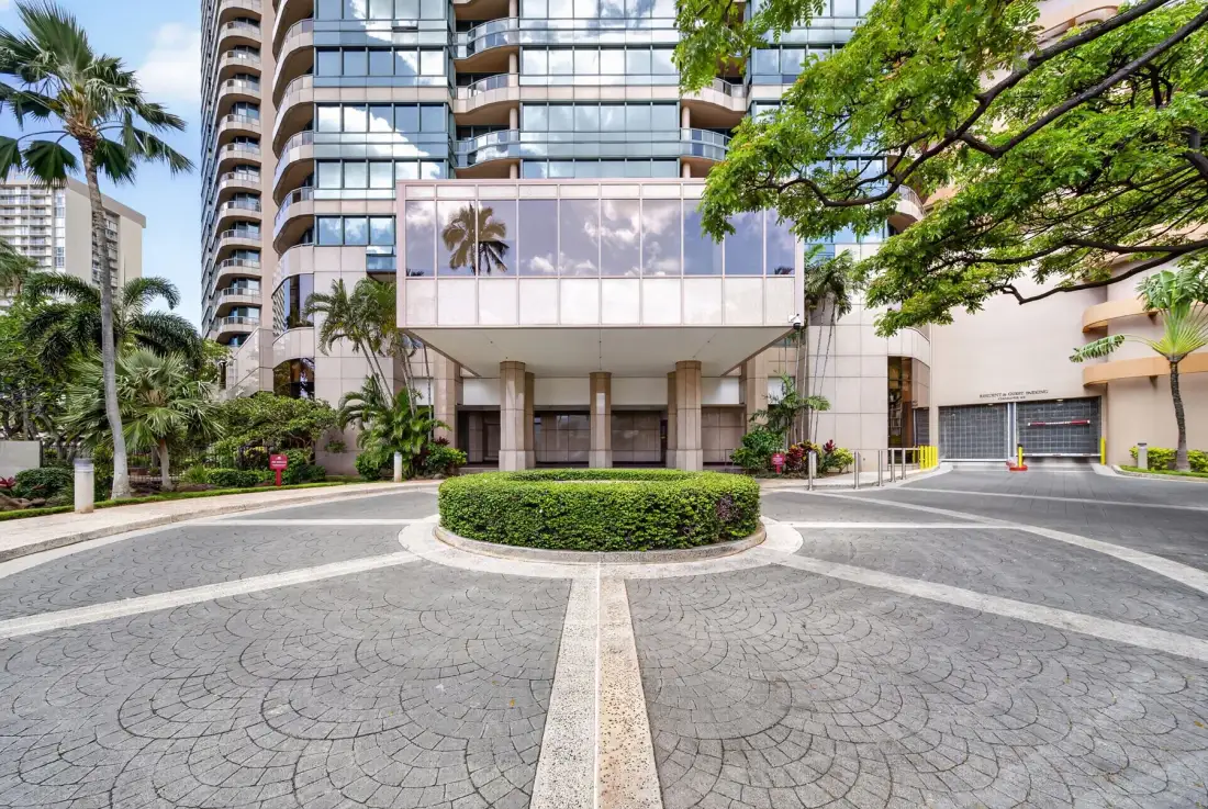 A high-rise building entrance faces a circular paved driveway and landscaped greenery.