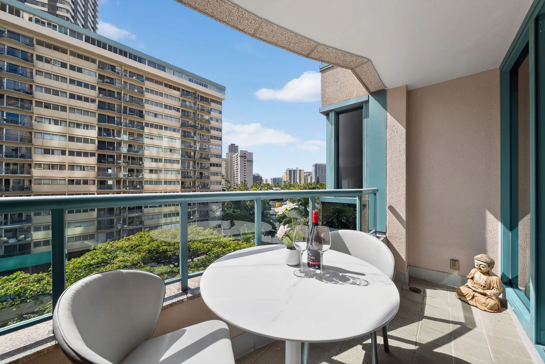 A balcony with a round table and seating overlooks nearby buildings and greenery.