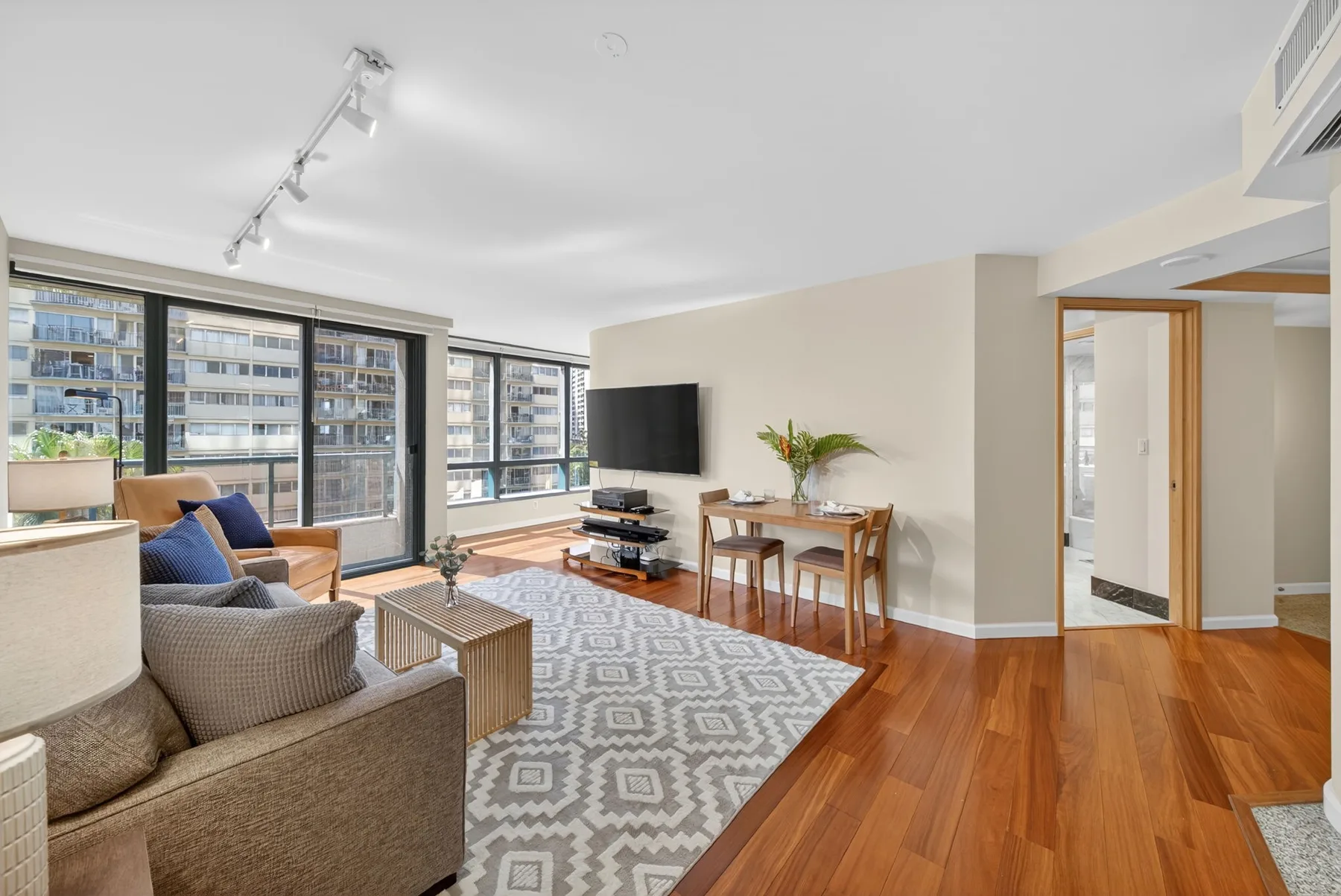 A bright living and dining area has large windows, seating, and a wooden floor.