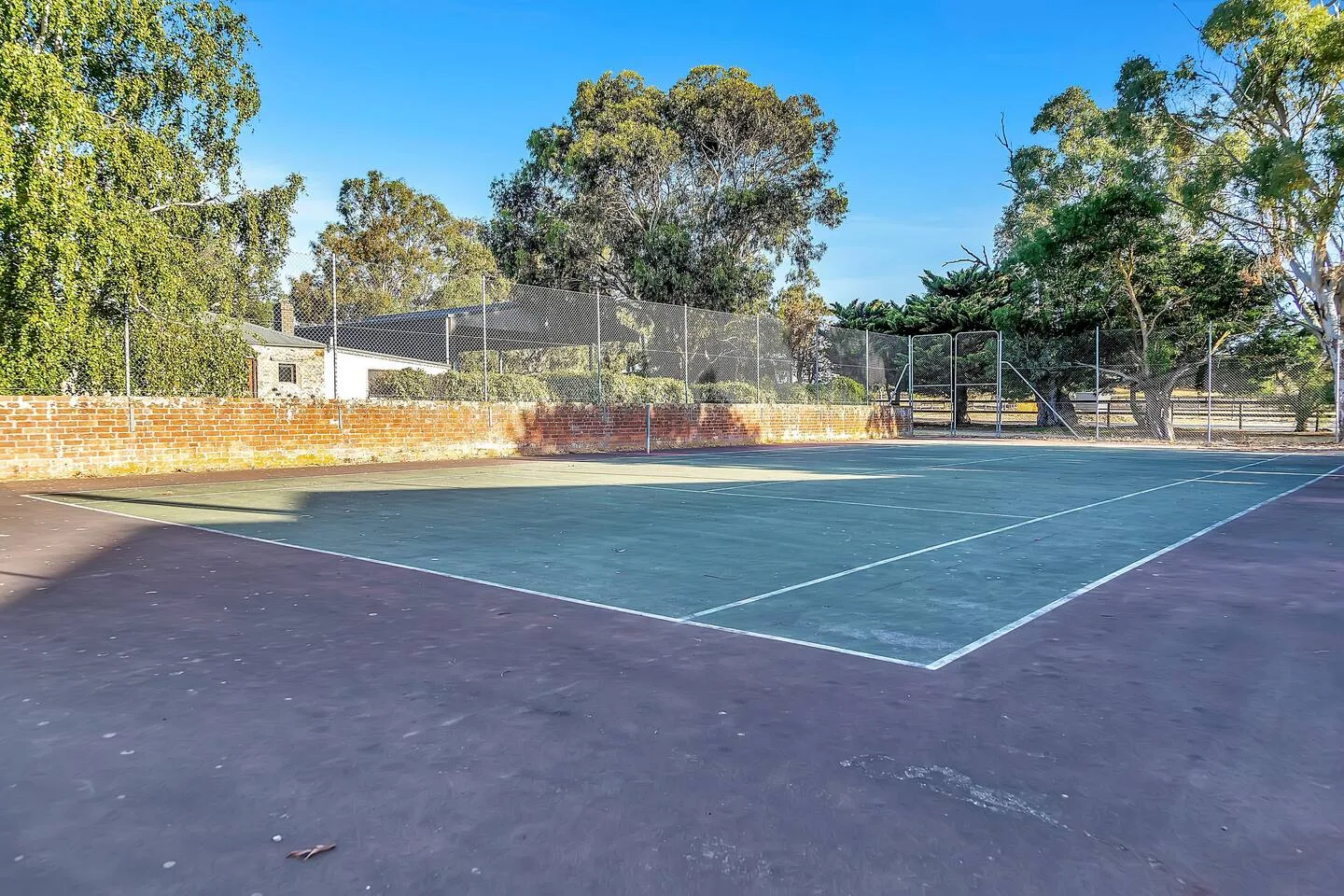 Una pista de tenis al aire libre está rodeada de árboles y un muro bajo de ladrillo.