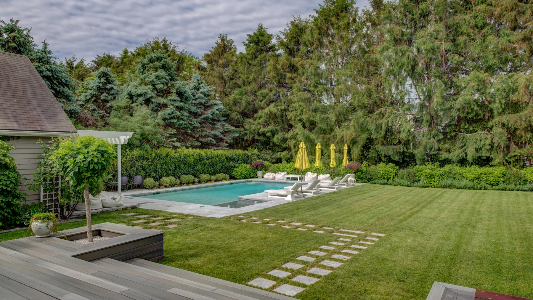 Un jardín trasero ajardinado tiene una piscina, césped y un camino de piedras.