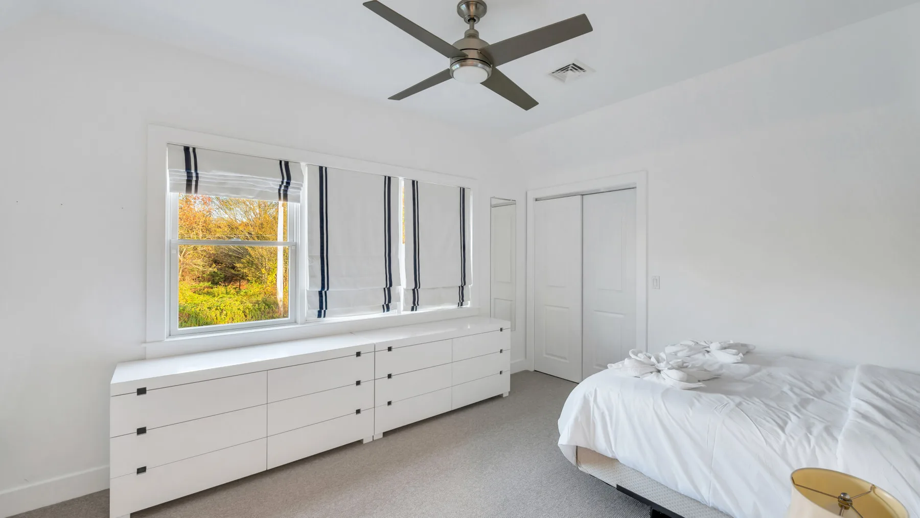 Un dormitorio luminoso con ropa de cama blanca, un ventilador de techo y ventanas con vista a los árboles.