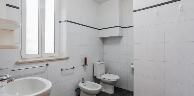 A tiled bathroom with a toilet, bidet, and sink beneath a window.