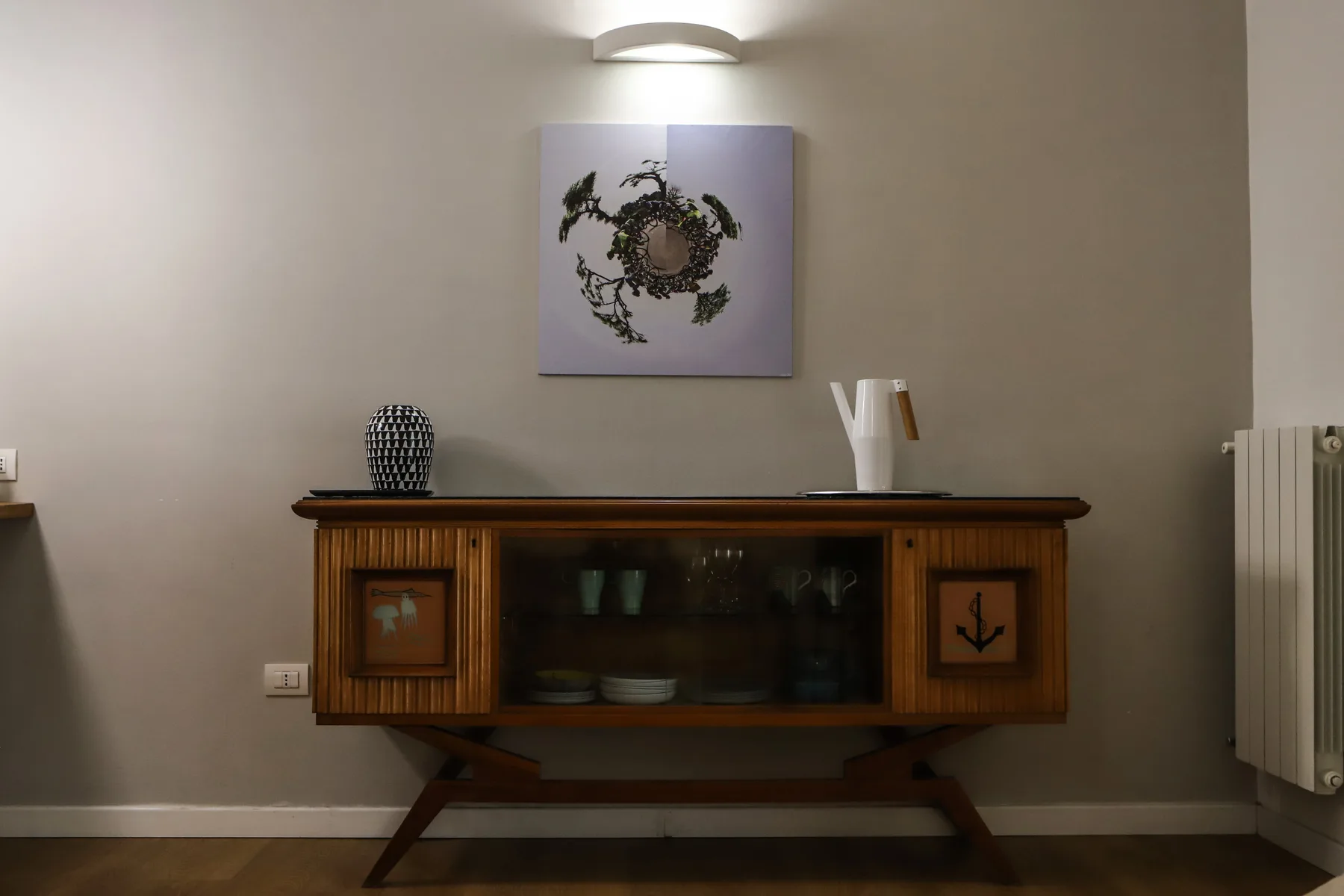 Un aparador de madera está bajo una obra botánica enmarcada en una habitación con luz suave.