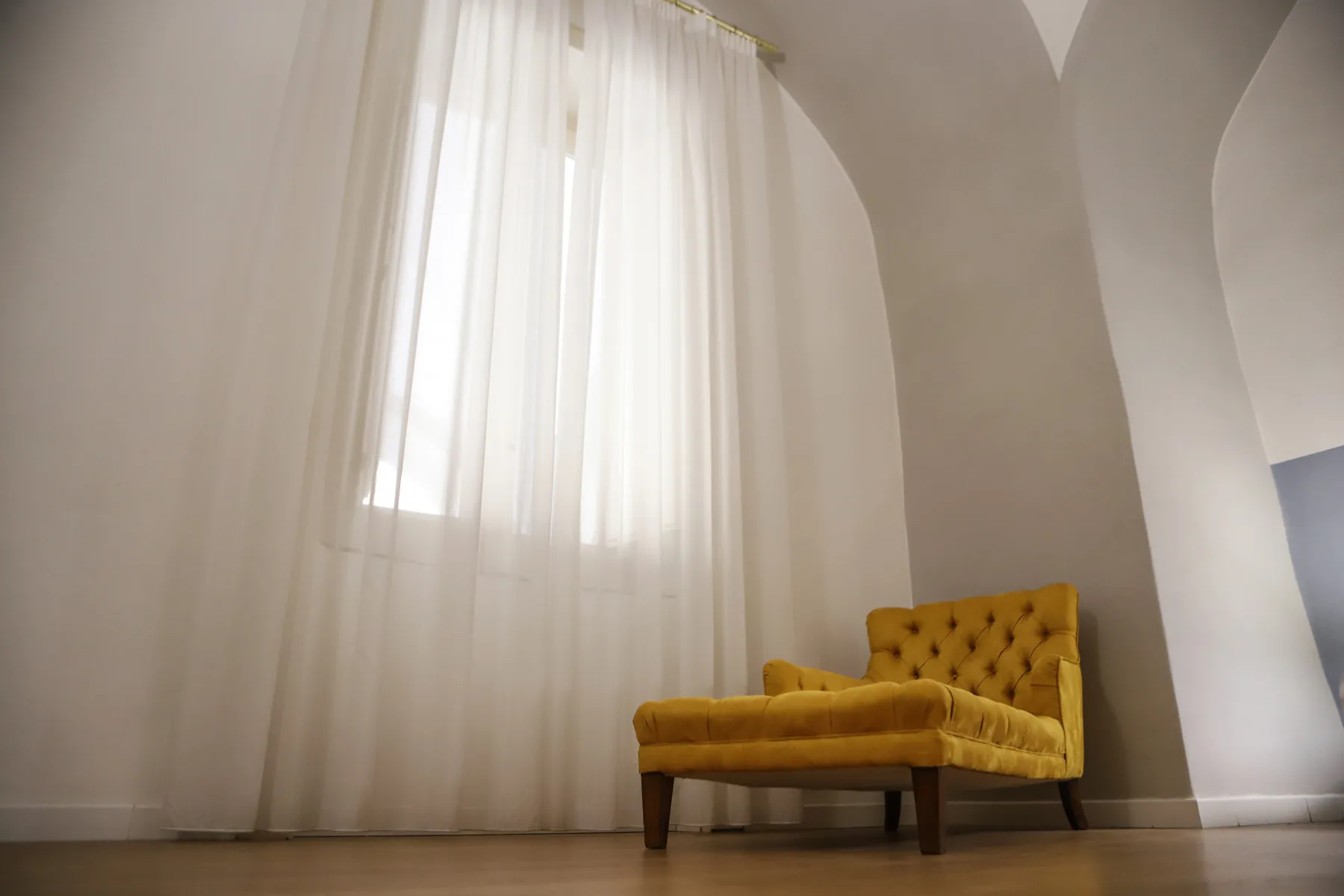 Un asiento tapizado color mostaza está bajo una ventana alta en arco con cortinas translúcidas.