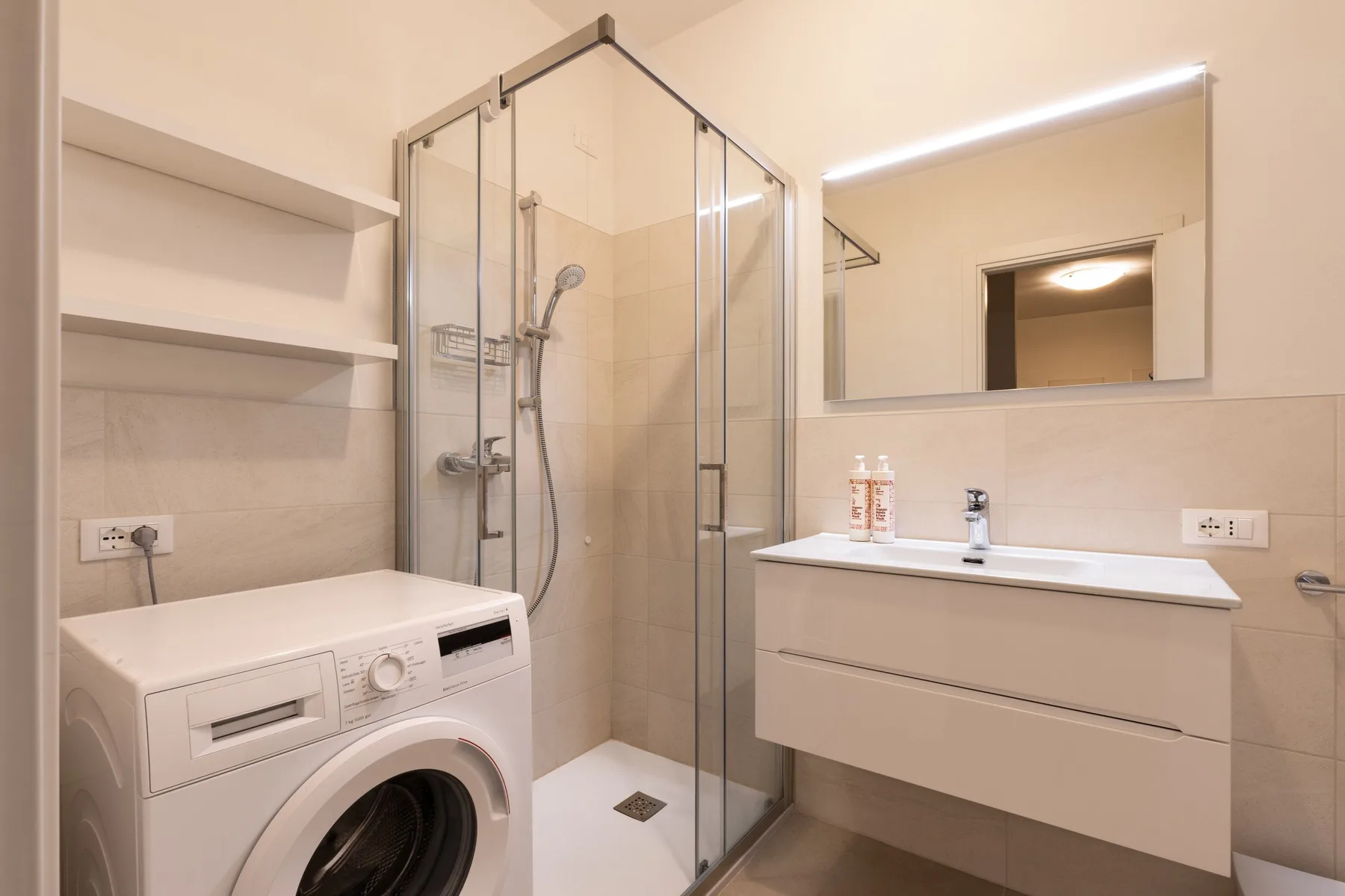 A bathroom with a glass shower, washing machine, and floating sink cabinet.