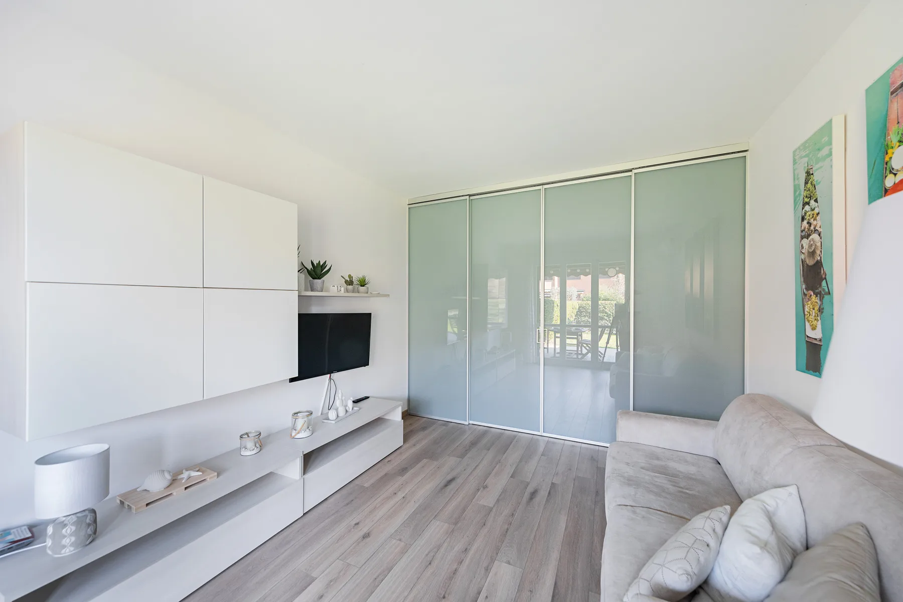 A bright living room with pale seating, a wall-mounted television, and sliding glass panels.