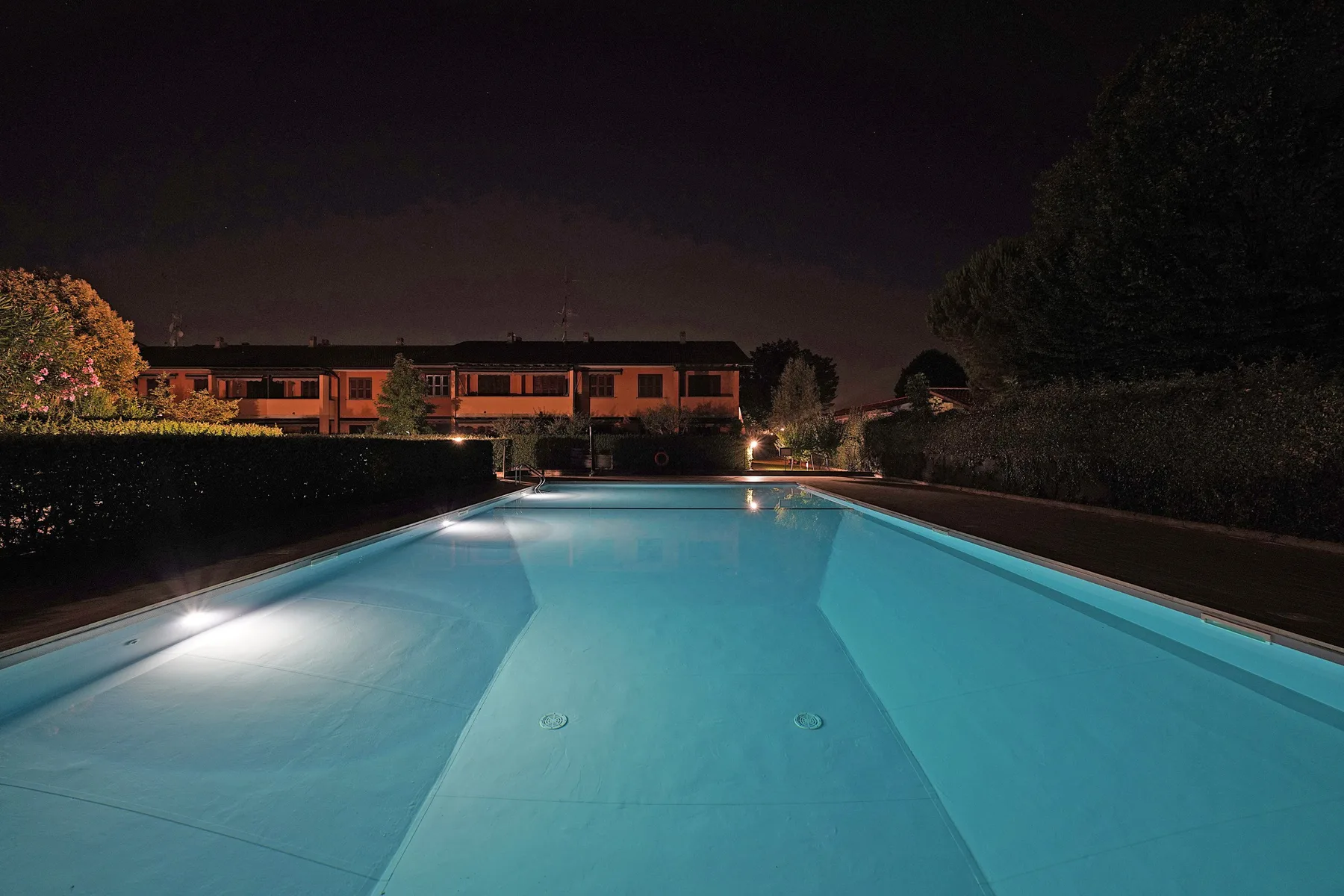 A swimming pool glows under nighttime lighting, with a building and trees in the background.