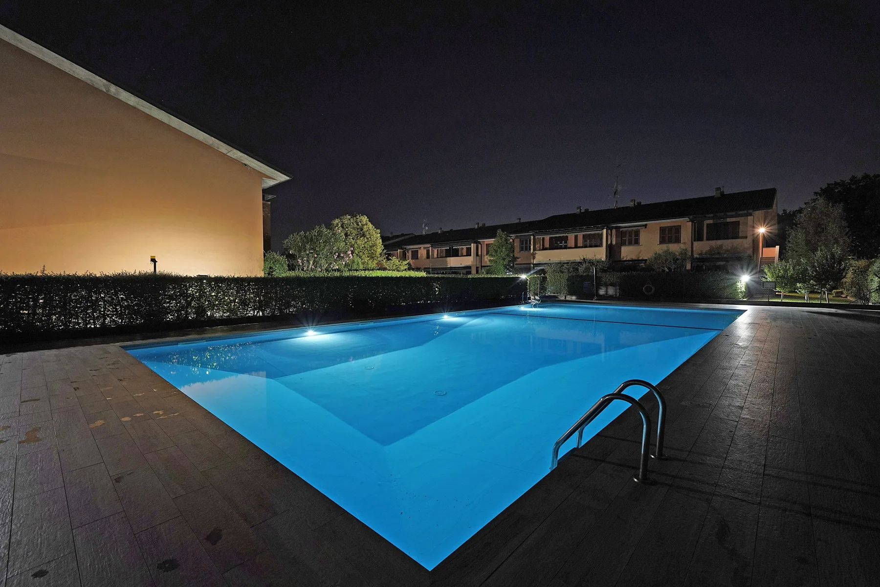 A swimming pool glows blue at night beside a paved deck.