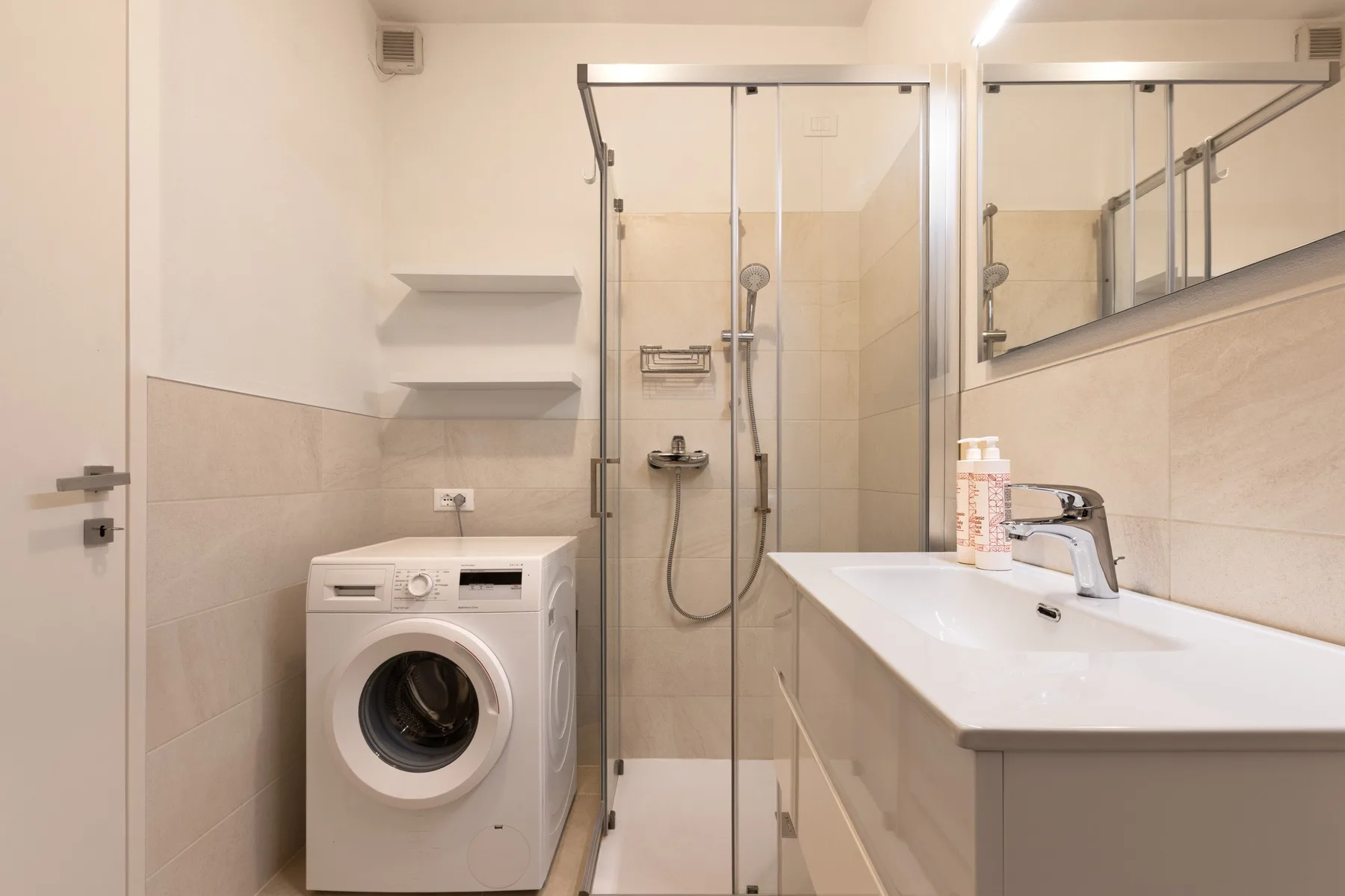 A bright bathroom with a glass shower, washing machine, and long vanity.