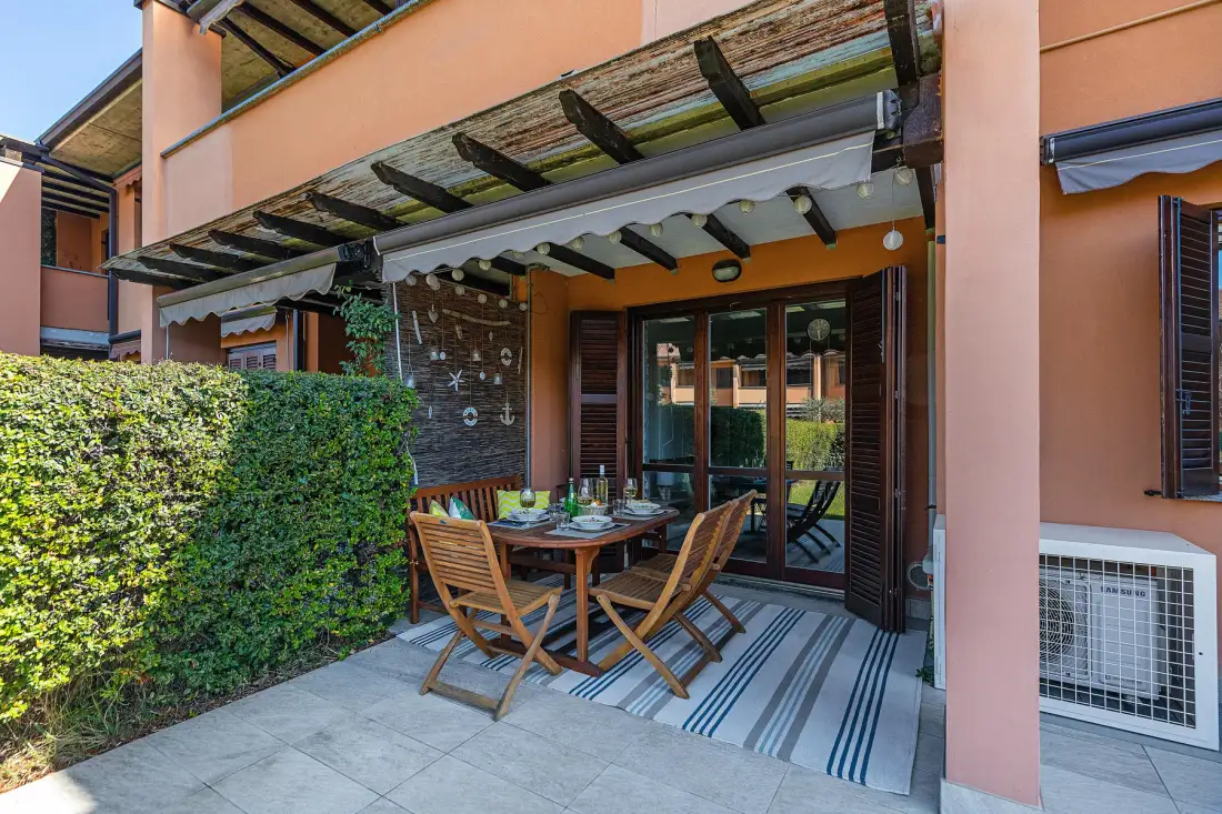 A covered patio has a dining table and wooden seating beside the house.