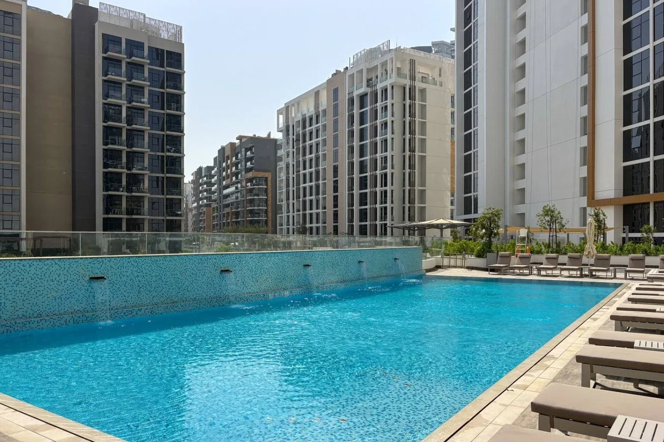 A swimming pool sits among modern apartment buildings, with lounge chairs along the deck.