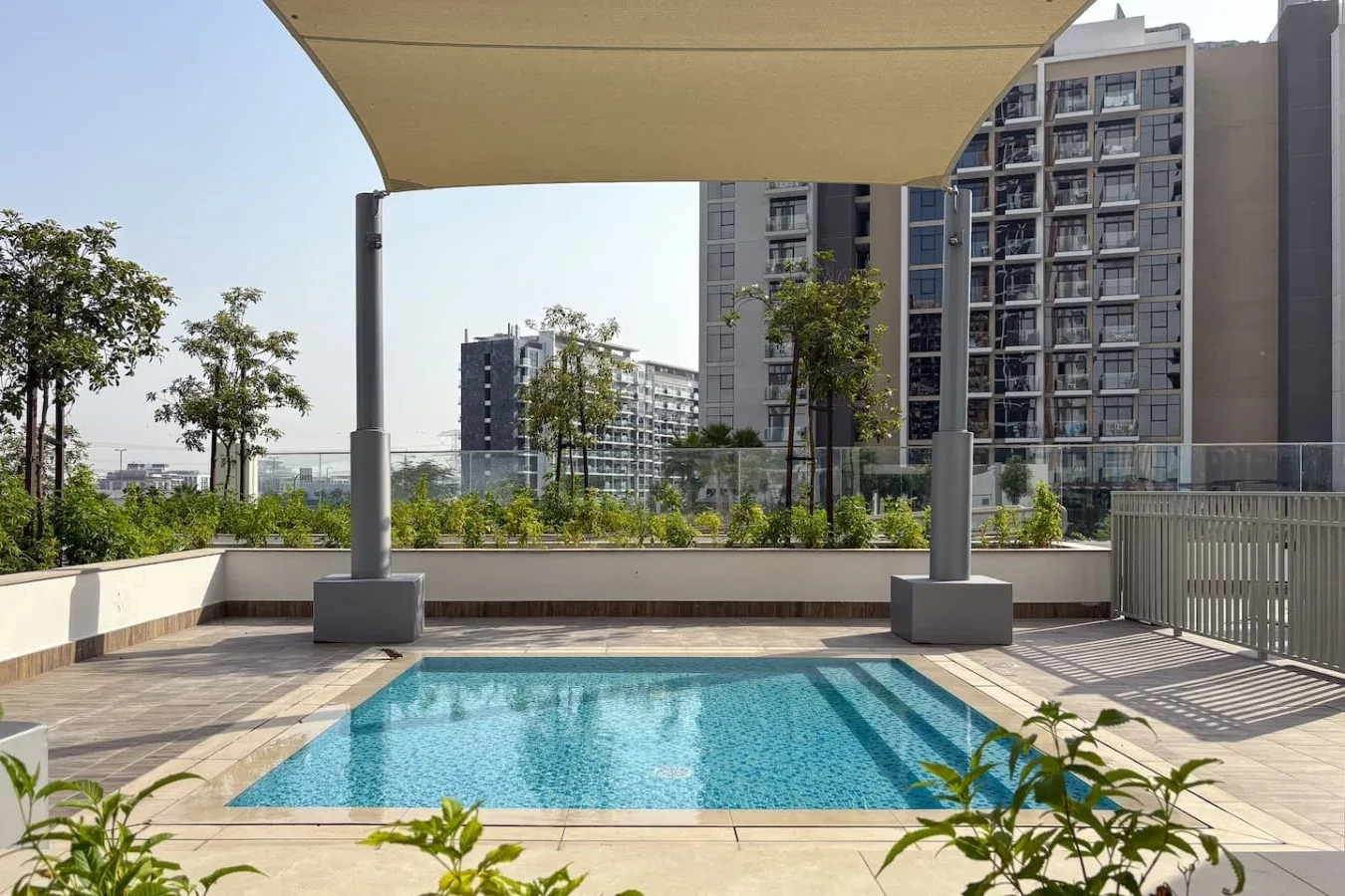 An outdoor swimming pool sits beneath a shade canopy, with apartment buildings in the background.