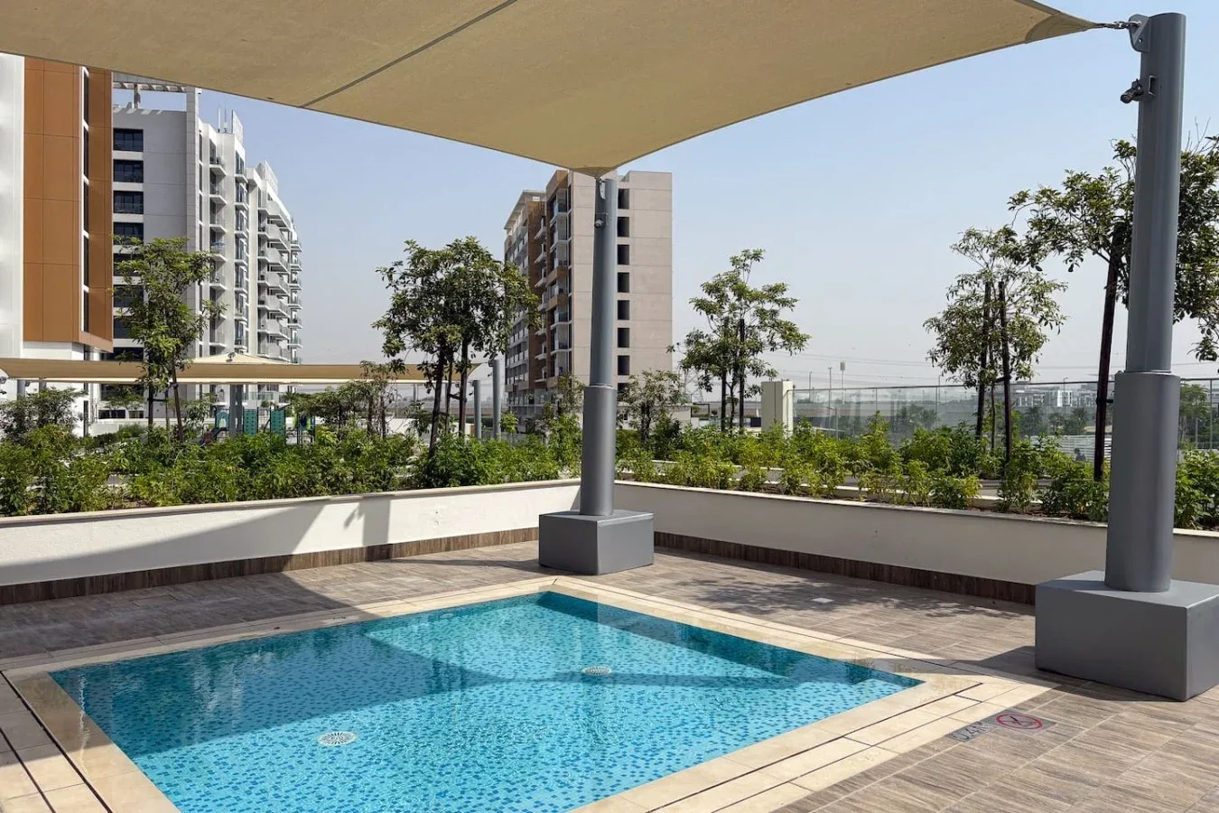 An outdoor swimming pool sits beneath a large shade canopy, with buildings and trees beyond.