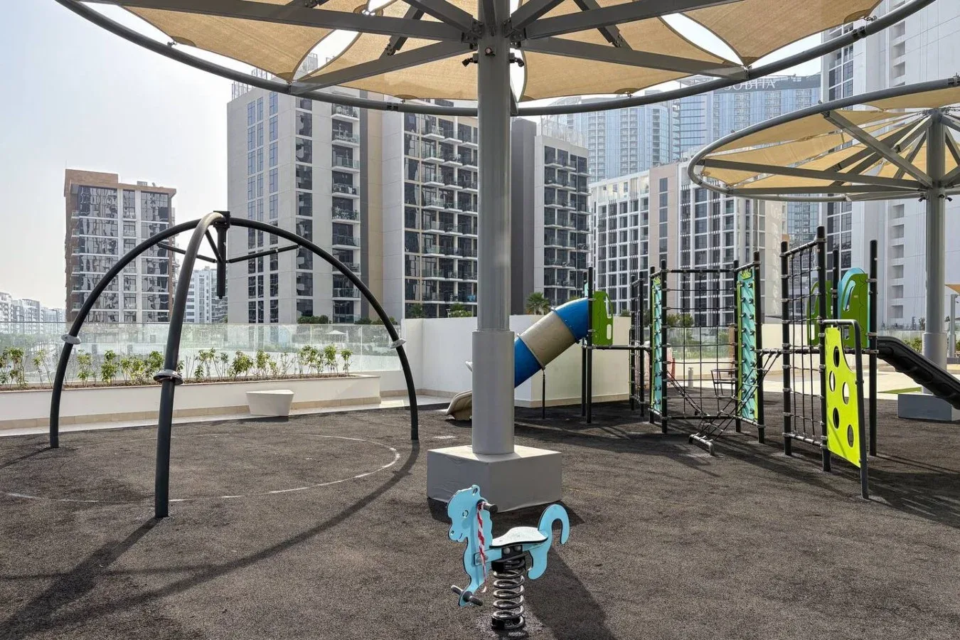 An outdoor playground with climbing equipment and shade canopies, surrounded by high-rise buildings.