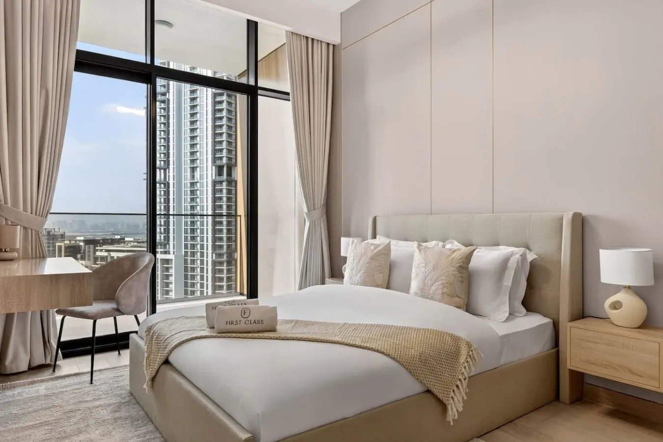 A bedroom with a neatly made bed beside large windows overlooking nearby buildings.